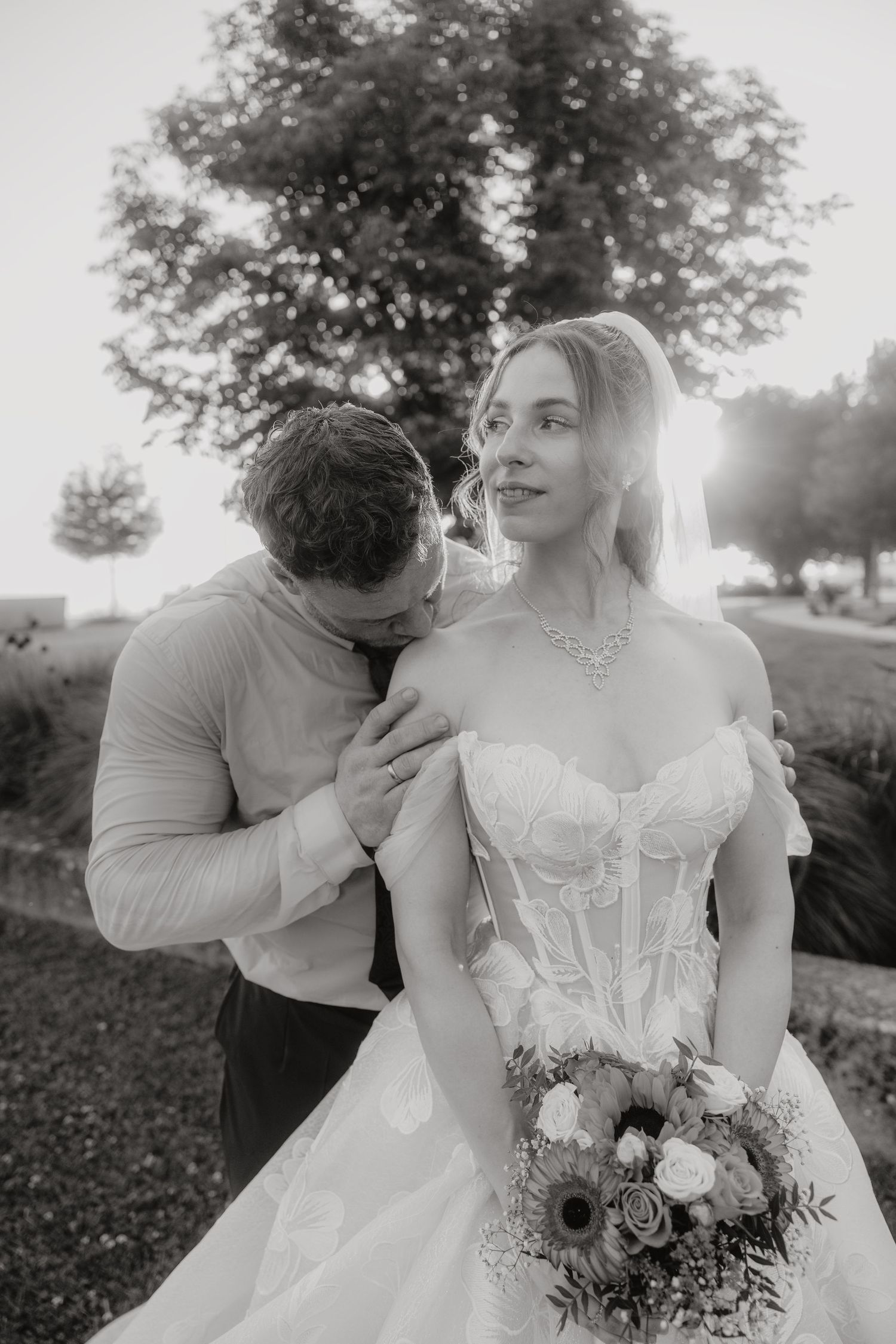Black and white photo of a groom kissing bride's shoulder as she holds a bouquet outdoors.