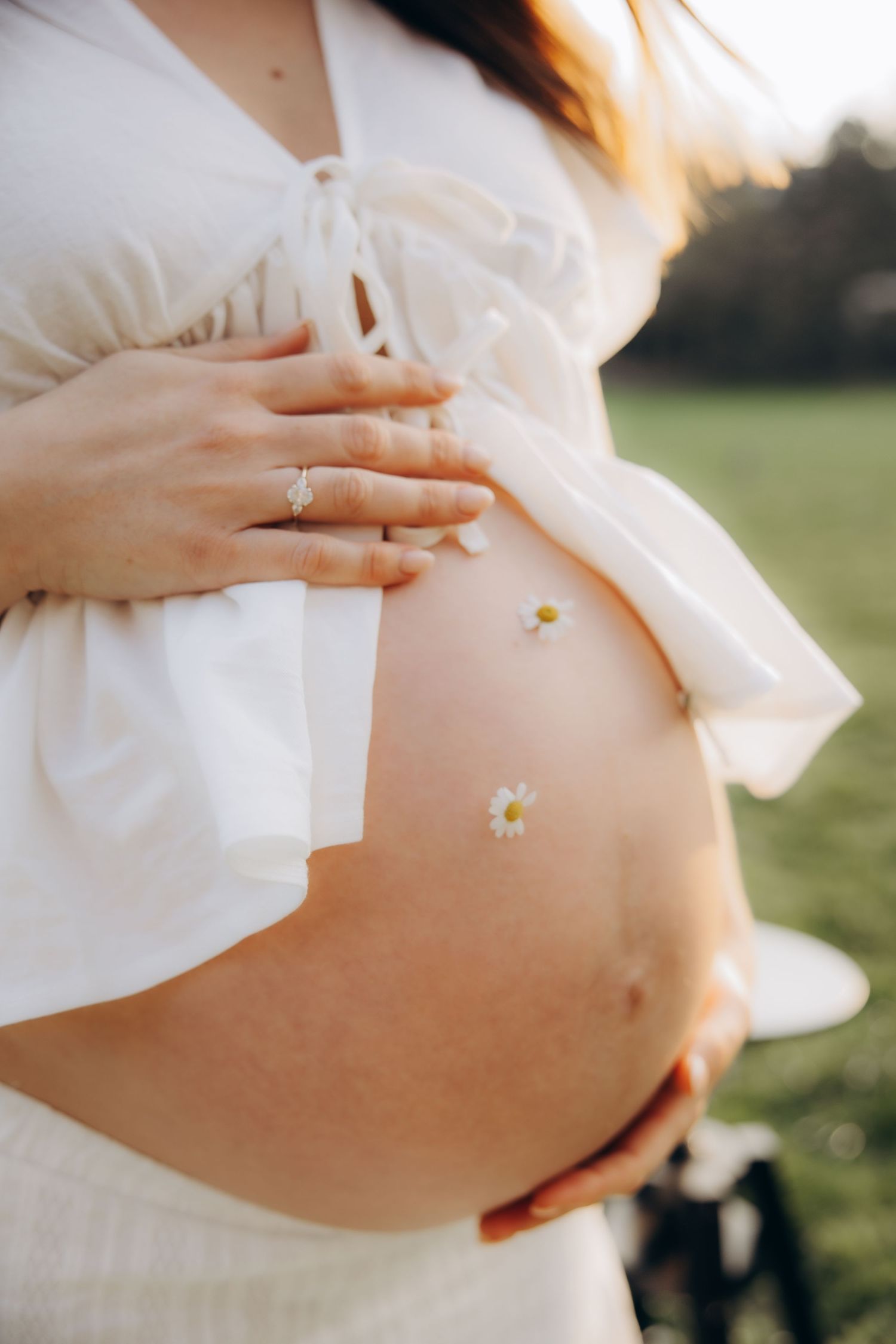 Close-up of a pregnant woman in white dress cradling her bare baby bump with daisy flowers outdoors.