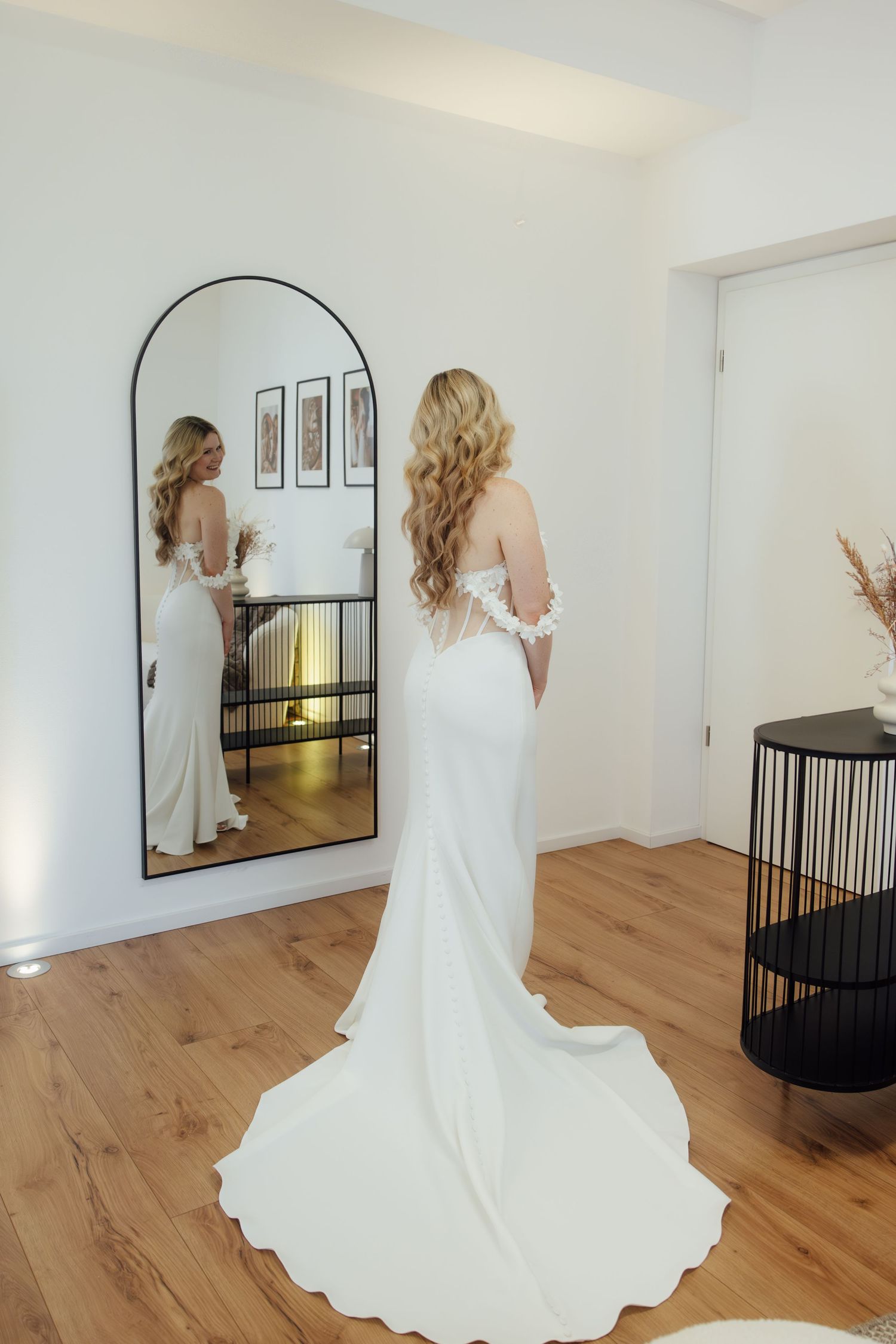 Bride in elegant white off-shoulder gown with train admiring her reflection in a large arched mirror.