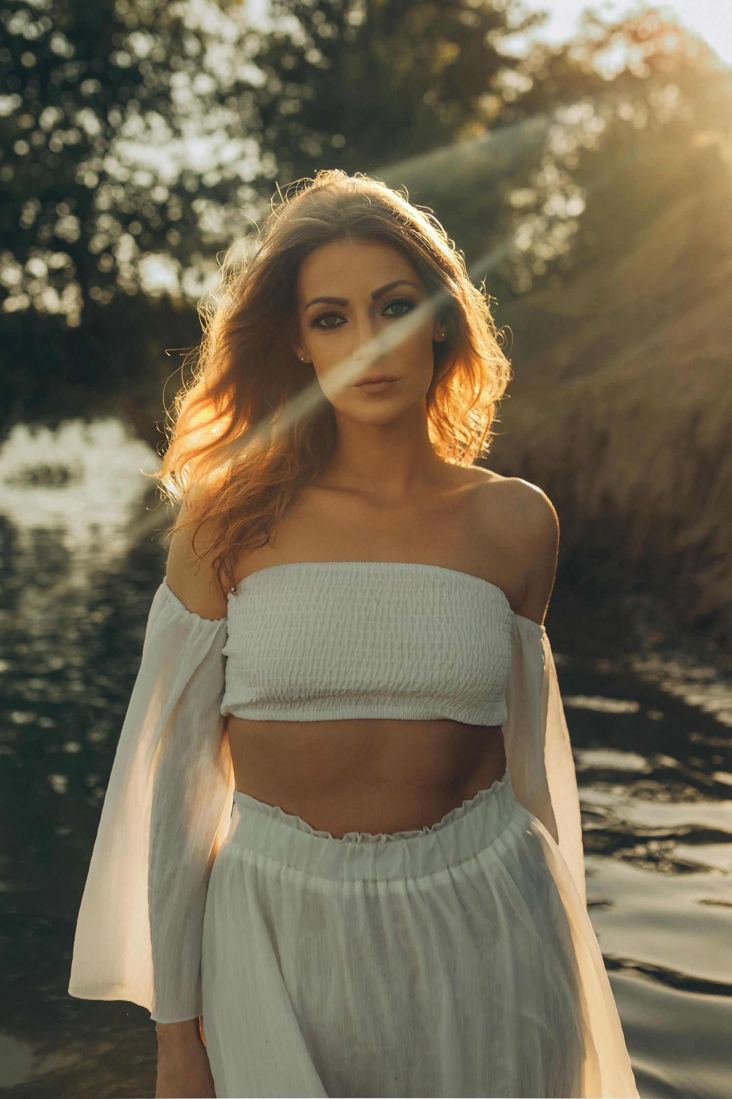 A woman in a white off-shoulder crop top and skirt standing by water in golden sunset light.
