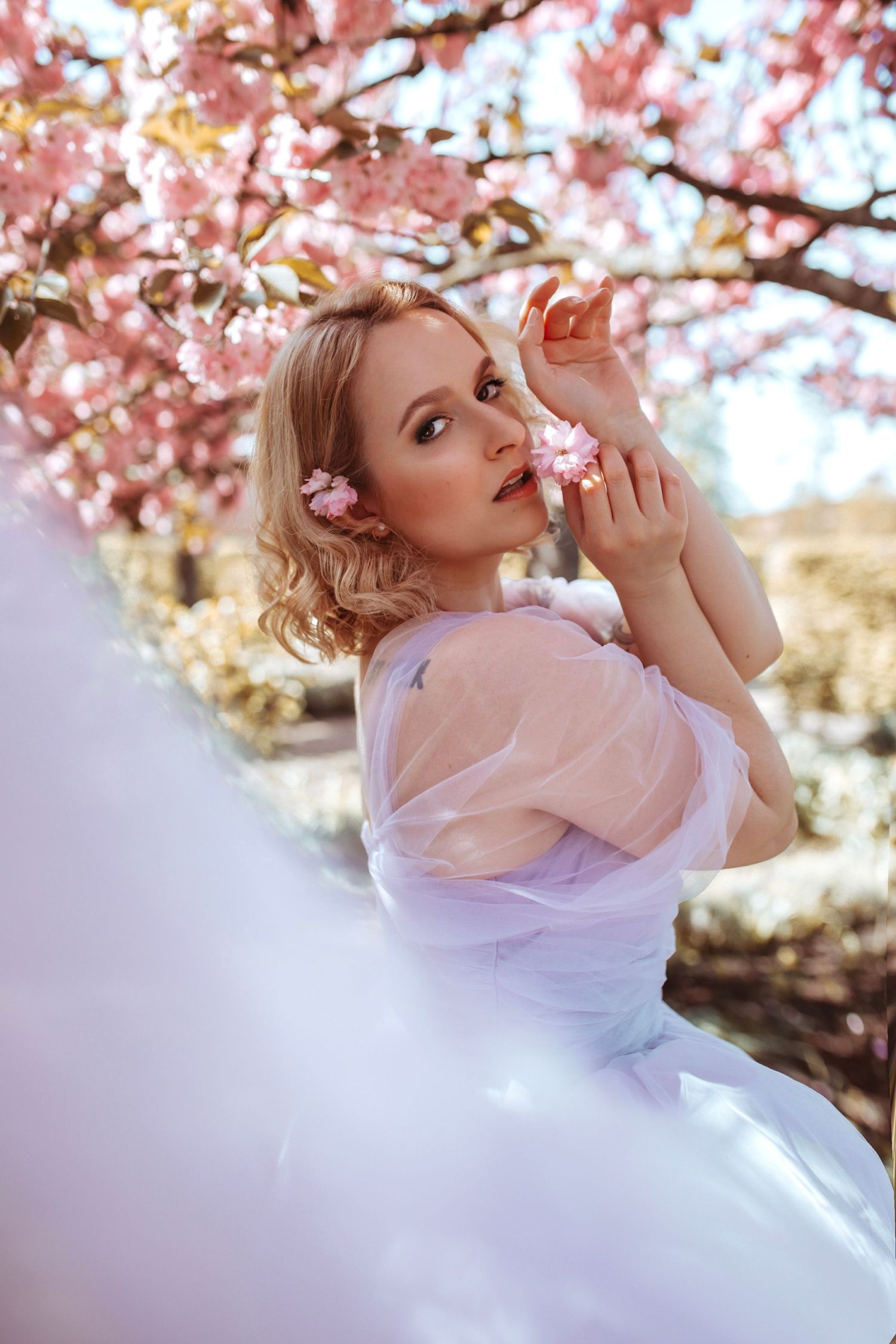 Young blonde woman in a white tulle dress posing near pink cherry blossom trees in spring.