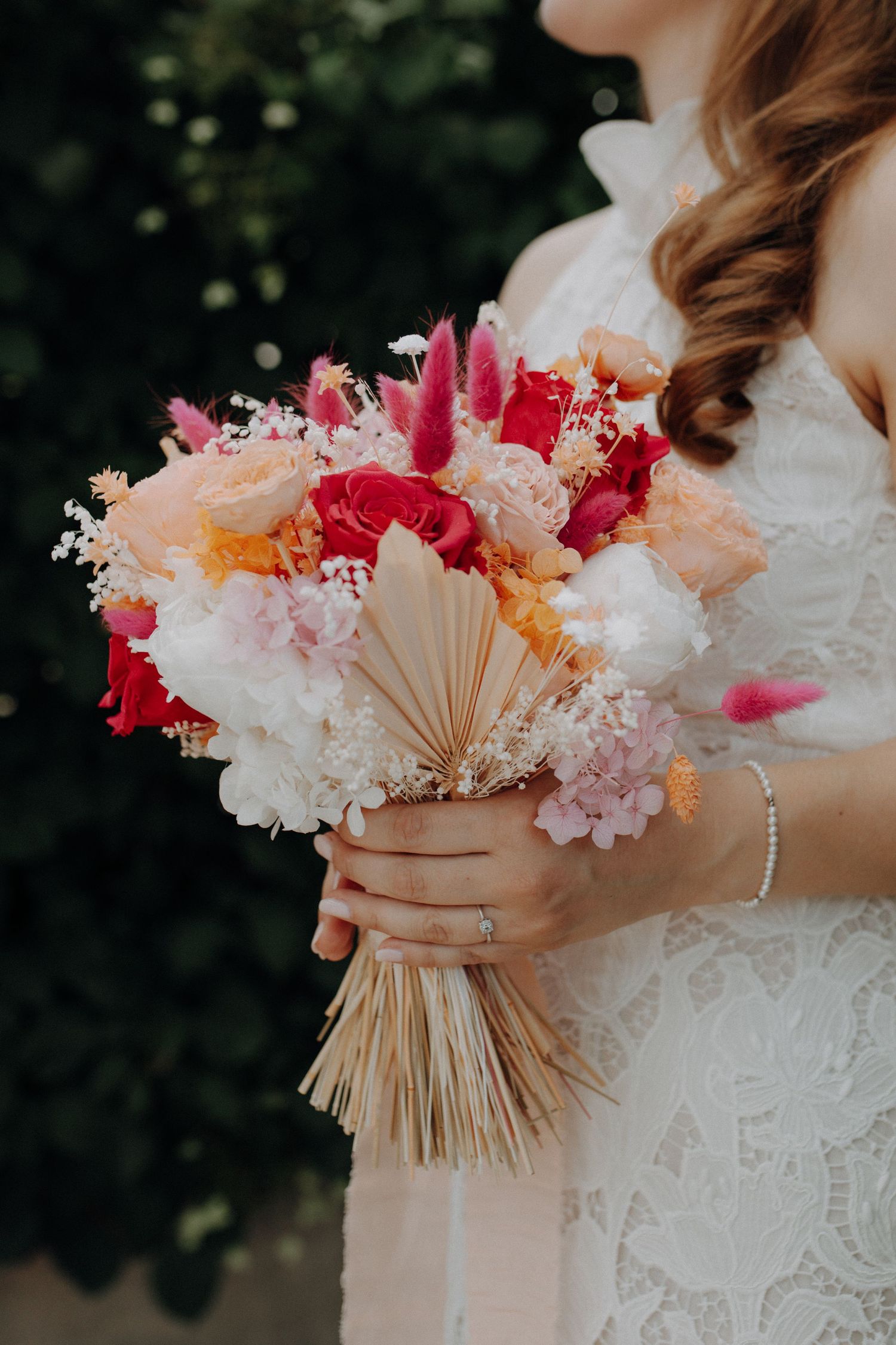 Bride holding a vibrant wedding bouquet with red, pink, orange, and white flowers with dried palm leaves.
