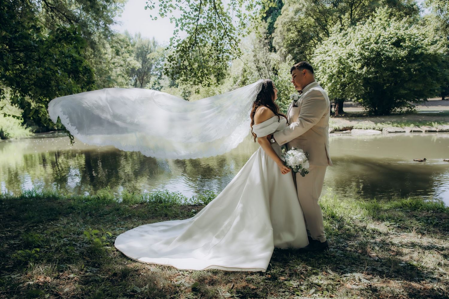 Bride in white gown with flowing veil and groom in tan suit embrace near a serene pond surrounded by lush green trees.