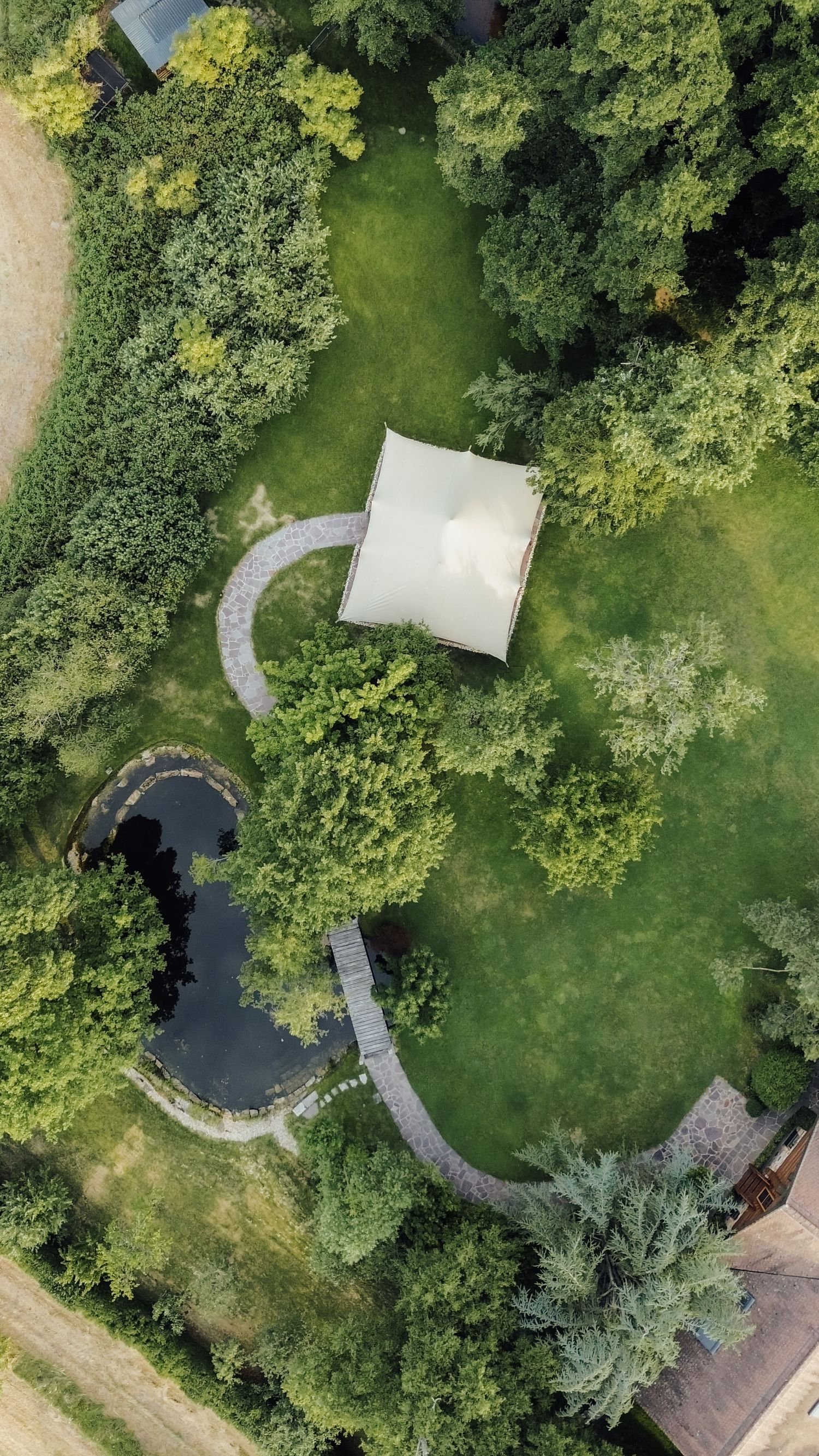 Aerial drone view of a white wedding tent surrounded by lush green trees and a small pond in a garden.