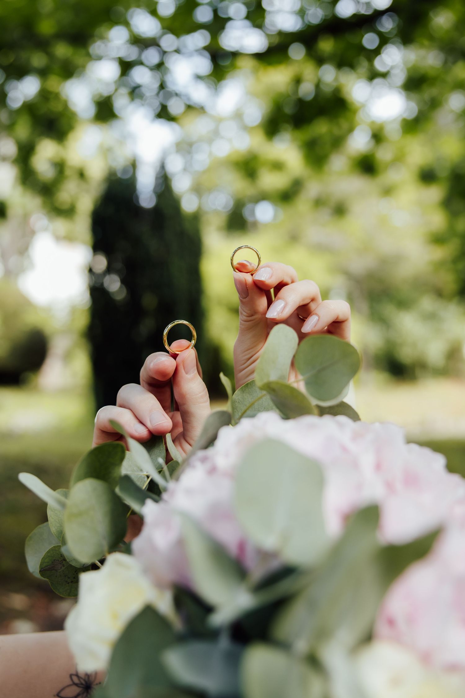 Two hands holding gold wedding rings above a bridal bouquet of pink hydrangeas and eucalyptus in a garden.