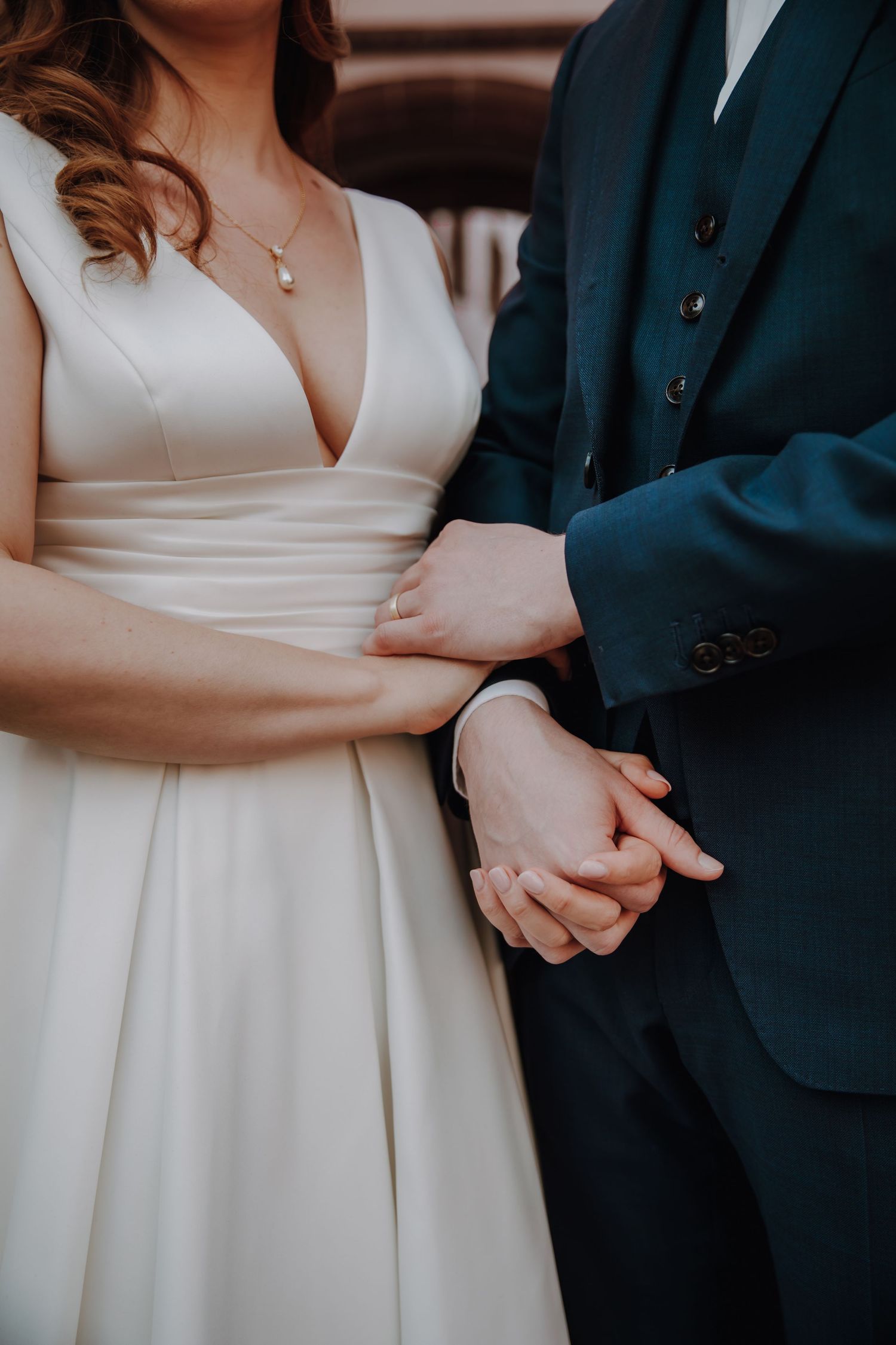 Bride in white gown and groom in navy suit holding hands, showcasing wedding rings during ceremony.