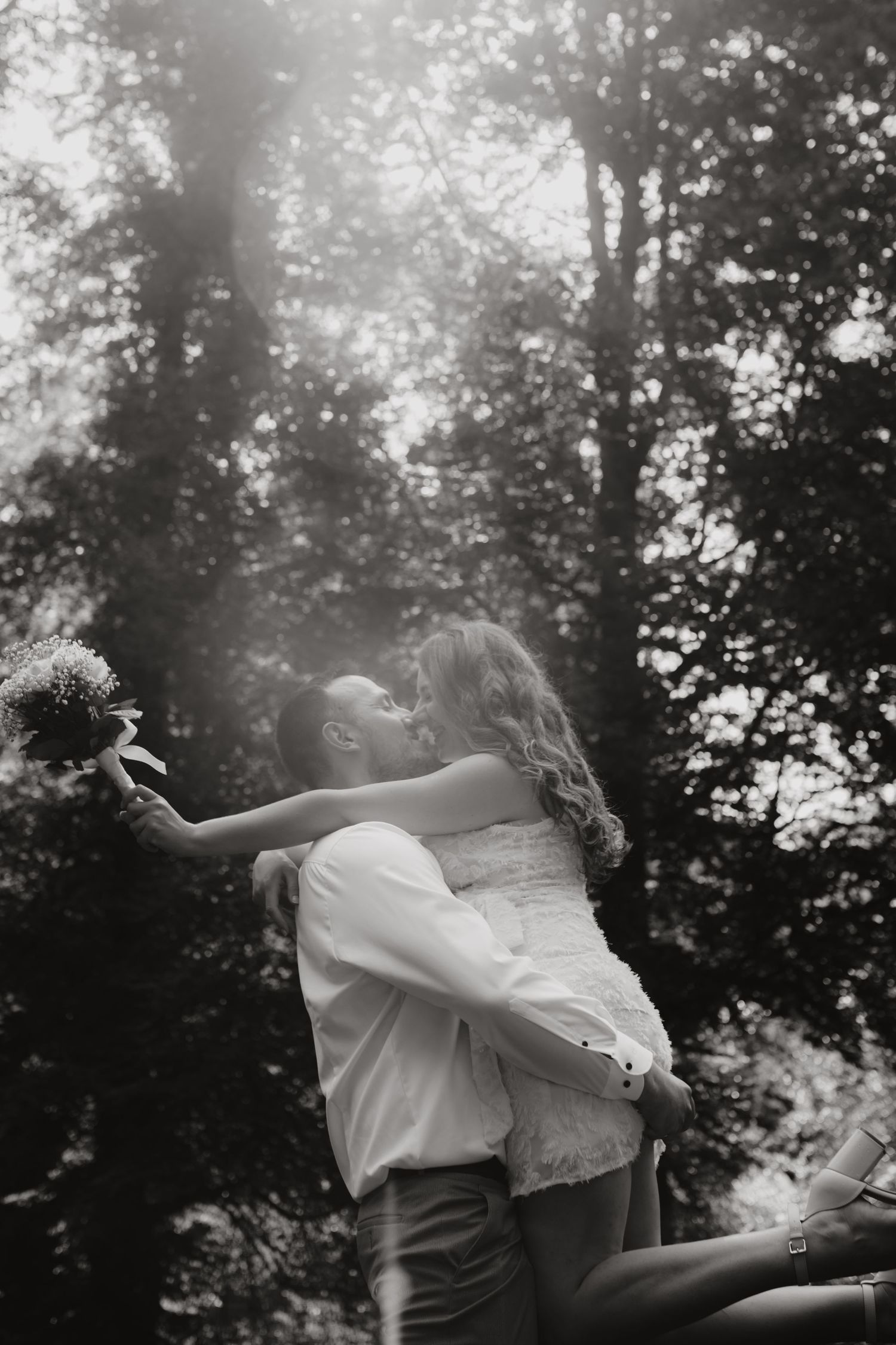 Black and white photo of bride and groom sharing a kiss in a sunlit forest setting.