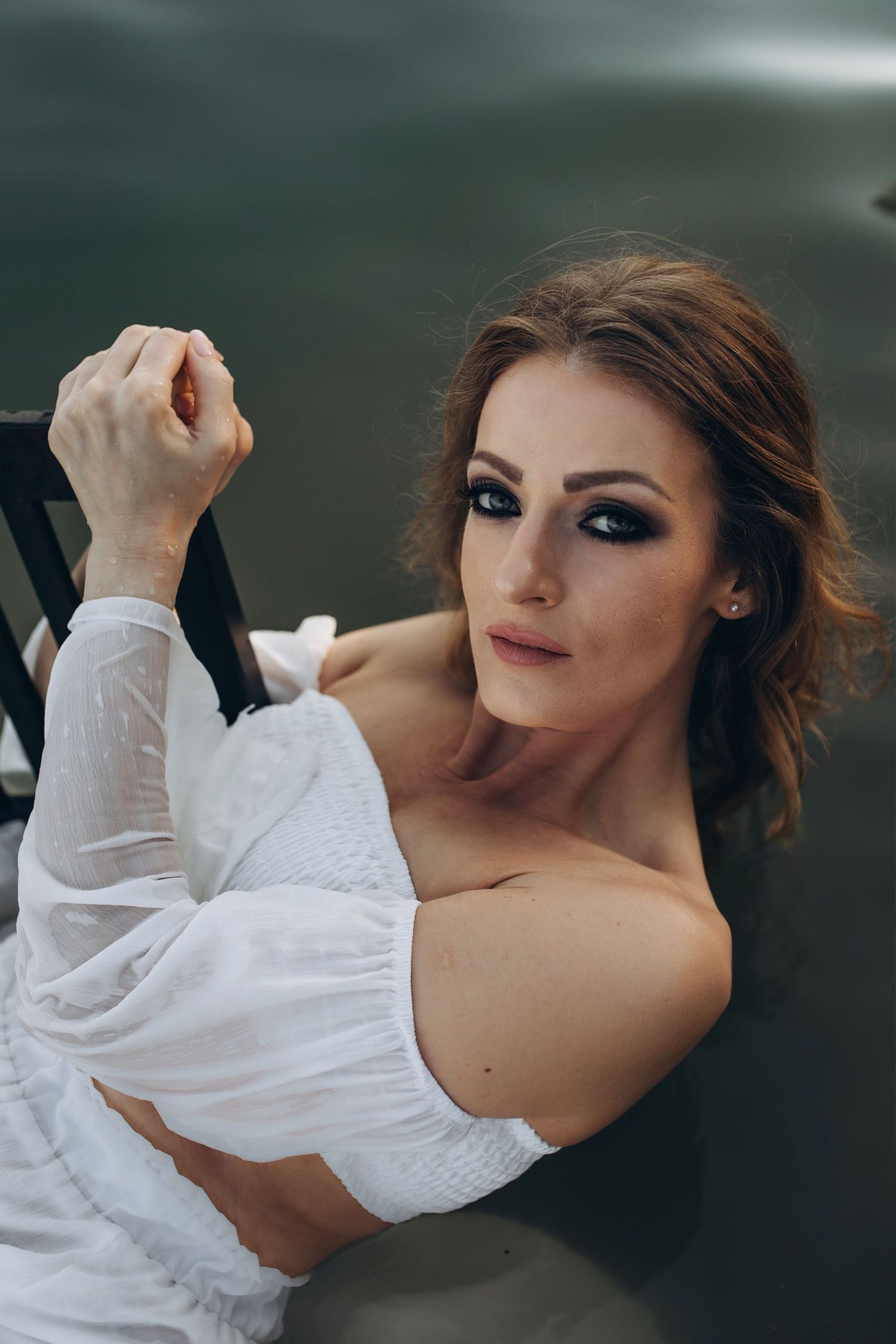 A woman in a white off-shoulder dress with dramatic eye makeup posing near dark water.