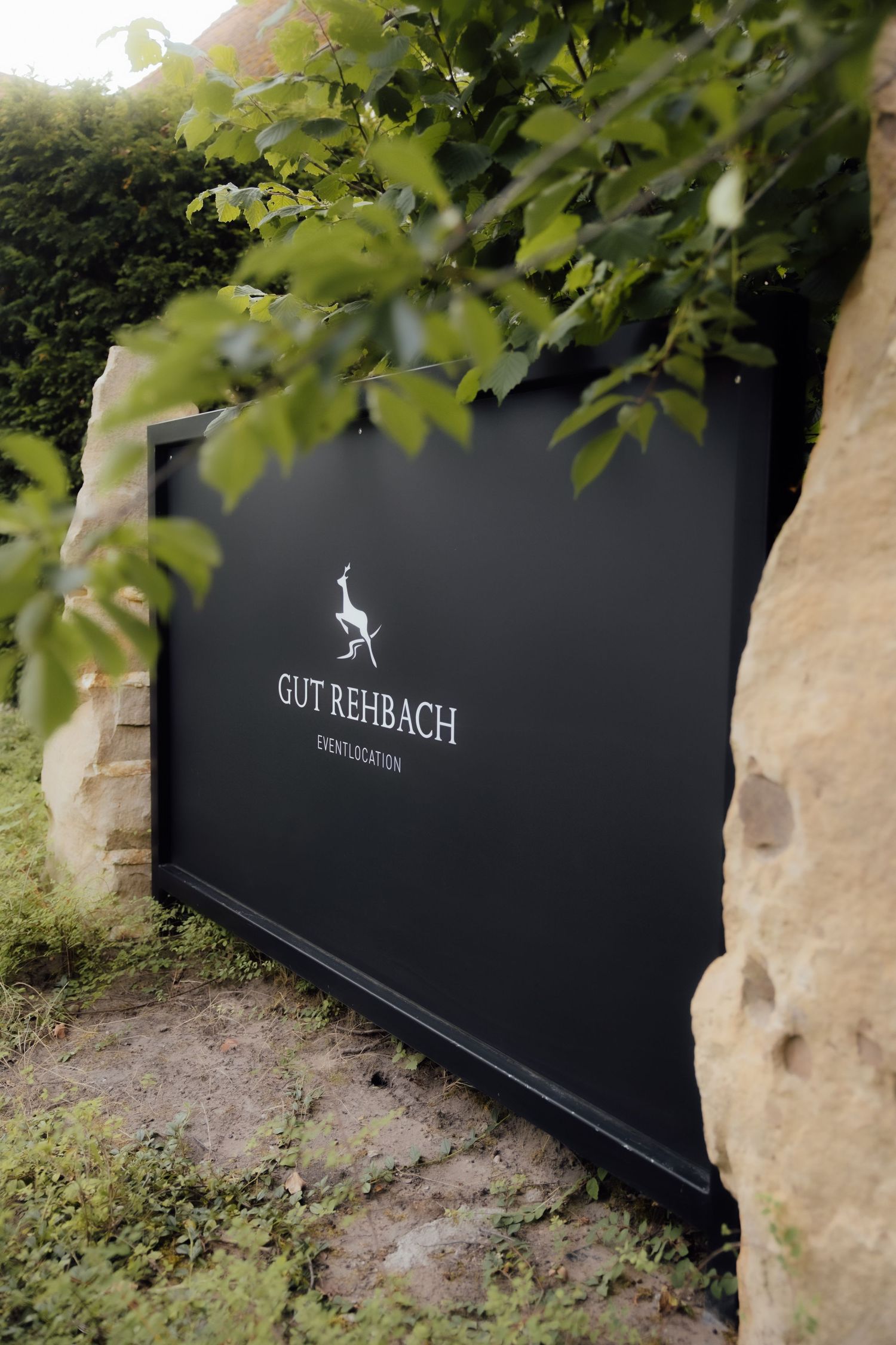 Black sign for Gut Rehbach Event Location surrounded by lush green foliage and rustic stone walls.