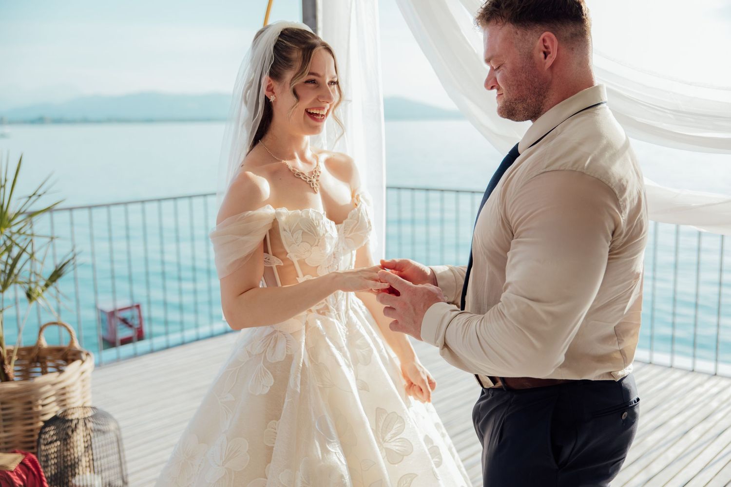 Groom placing wedding ring on bride's finger during ceremony with ocean view backdrop.