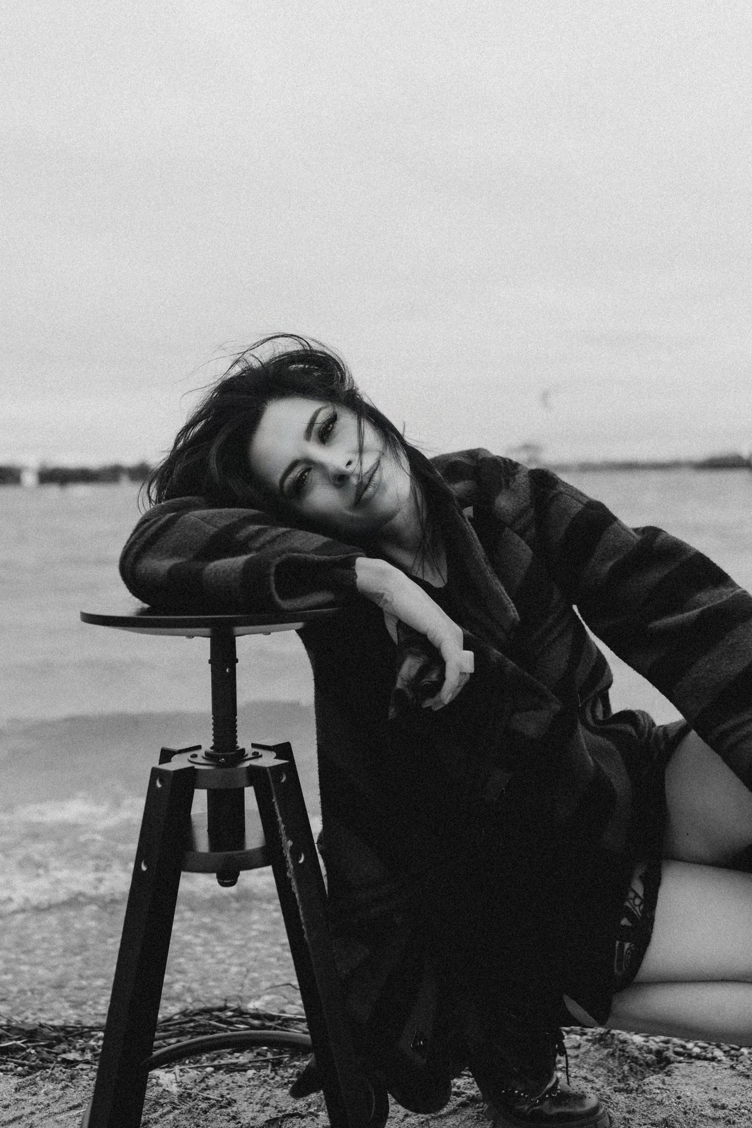 Black and white photo of a woman resting on a stool by the sea with windswept hair.