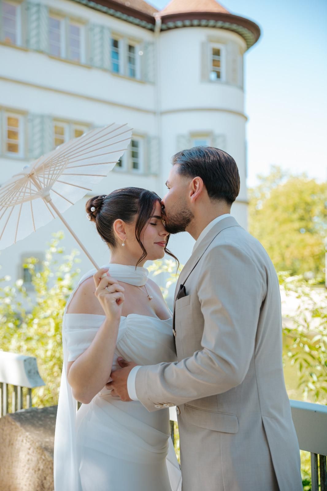 Bride and groom share a tender moment under a white parasol in front of an elegant manor.