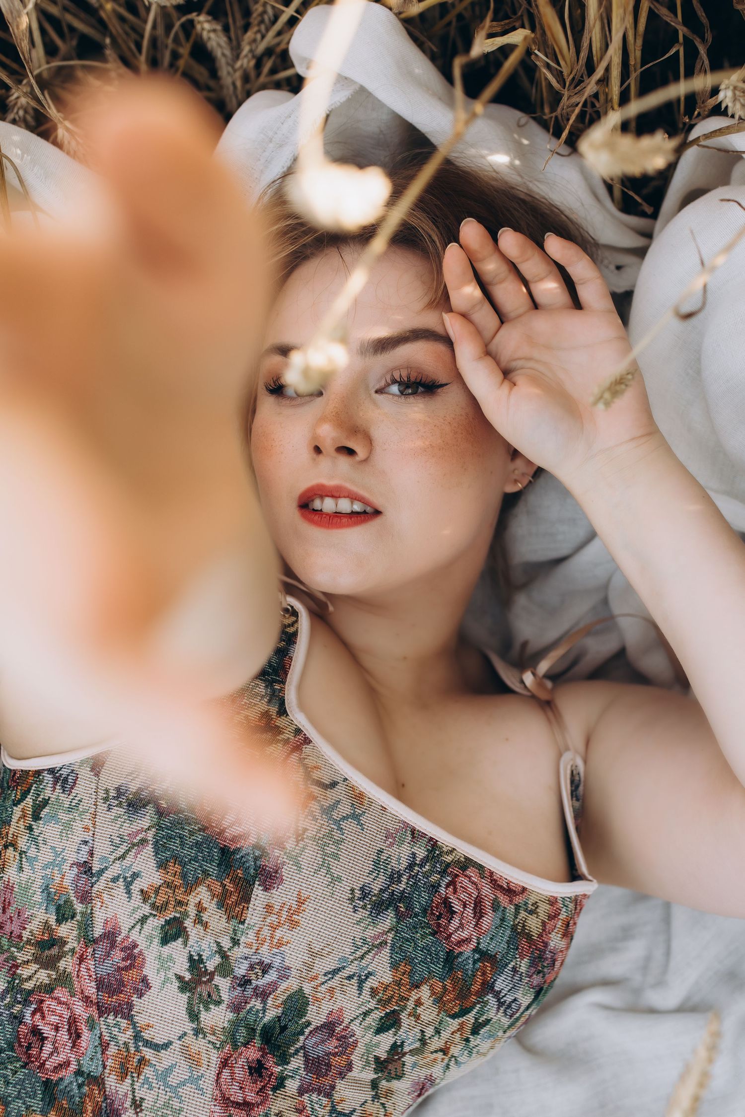Blonde woman with freckles and red lips lies in wheat field wearing floral dress, hand shielding eyes.