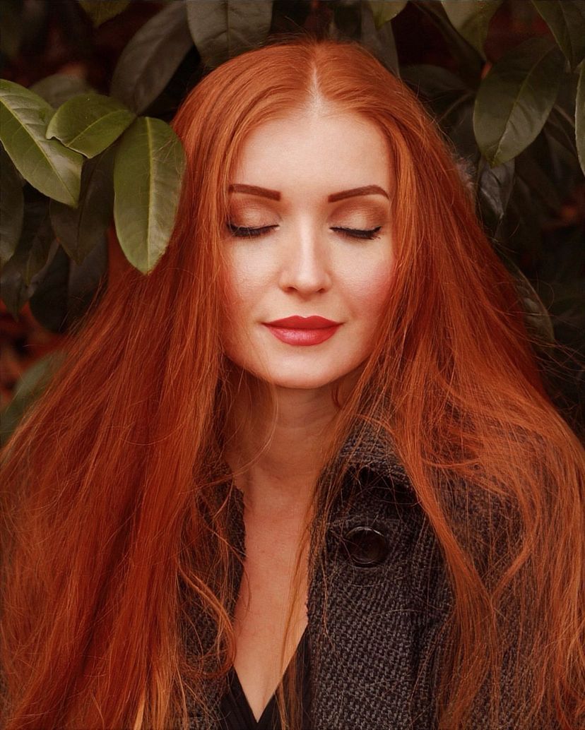 A woman with long red hair poses with closed eyes against a backdrop of green leaves.