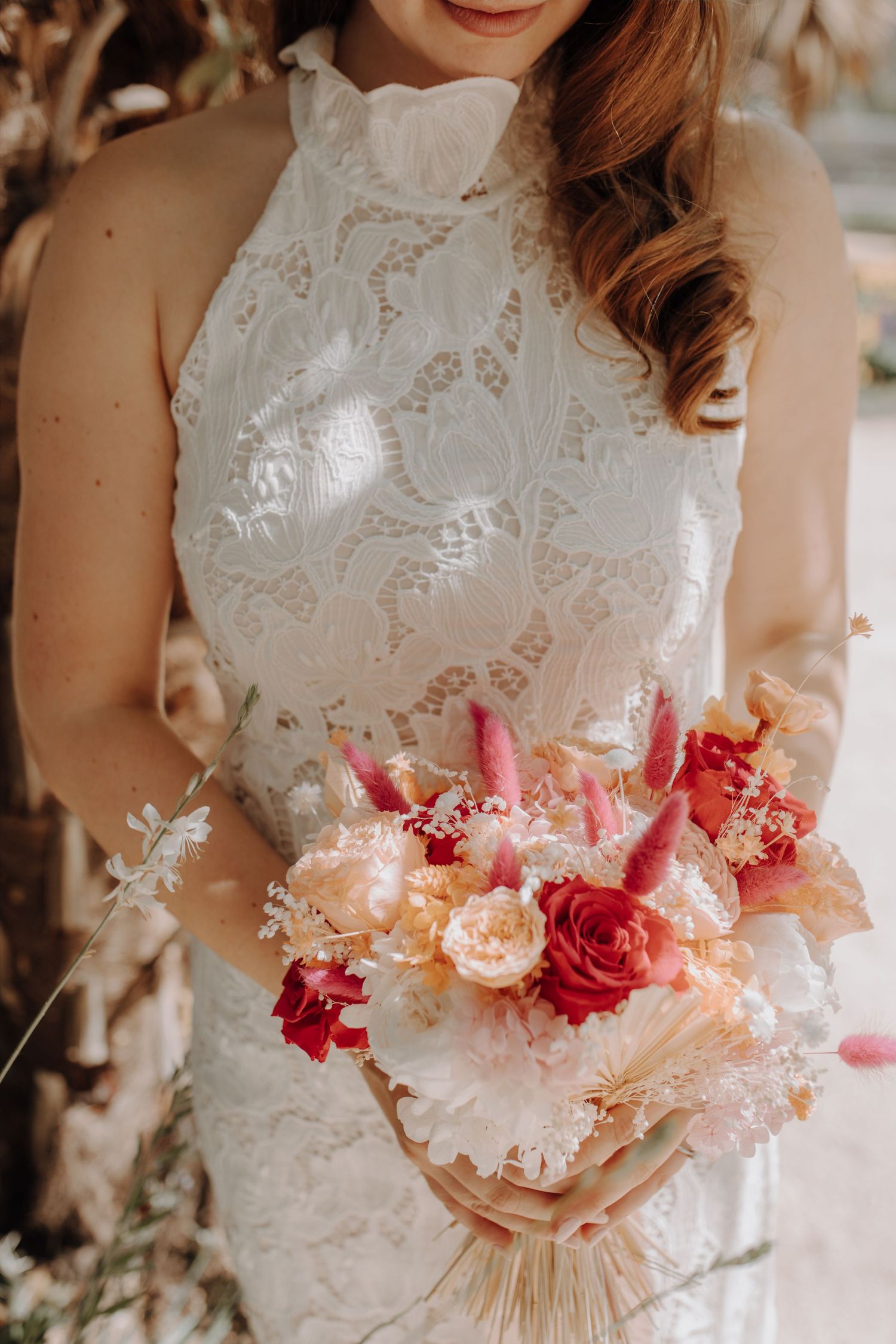 Bride in a lace halter wedding dress holding a colorful bouquet of red, pink, and peach flowers.