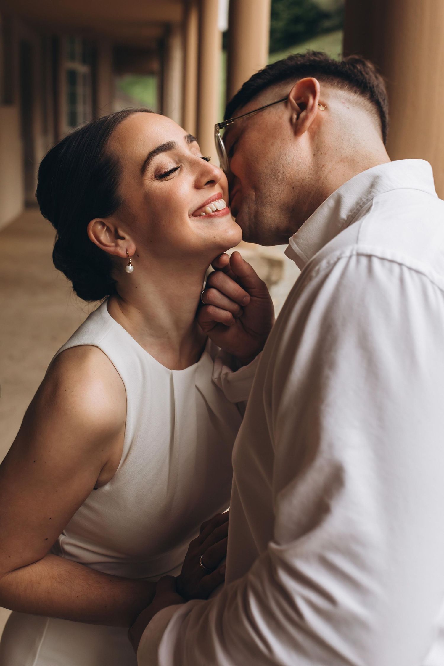 Happy bride in white dress smiling as groom kisses her cheek during romantic wedding moment outdoors.
