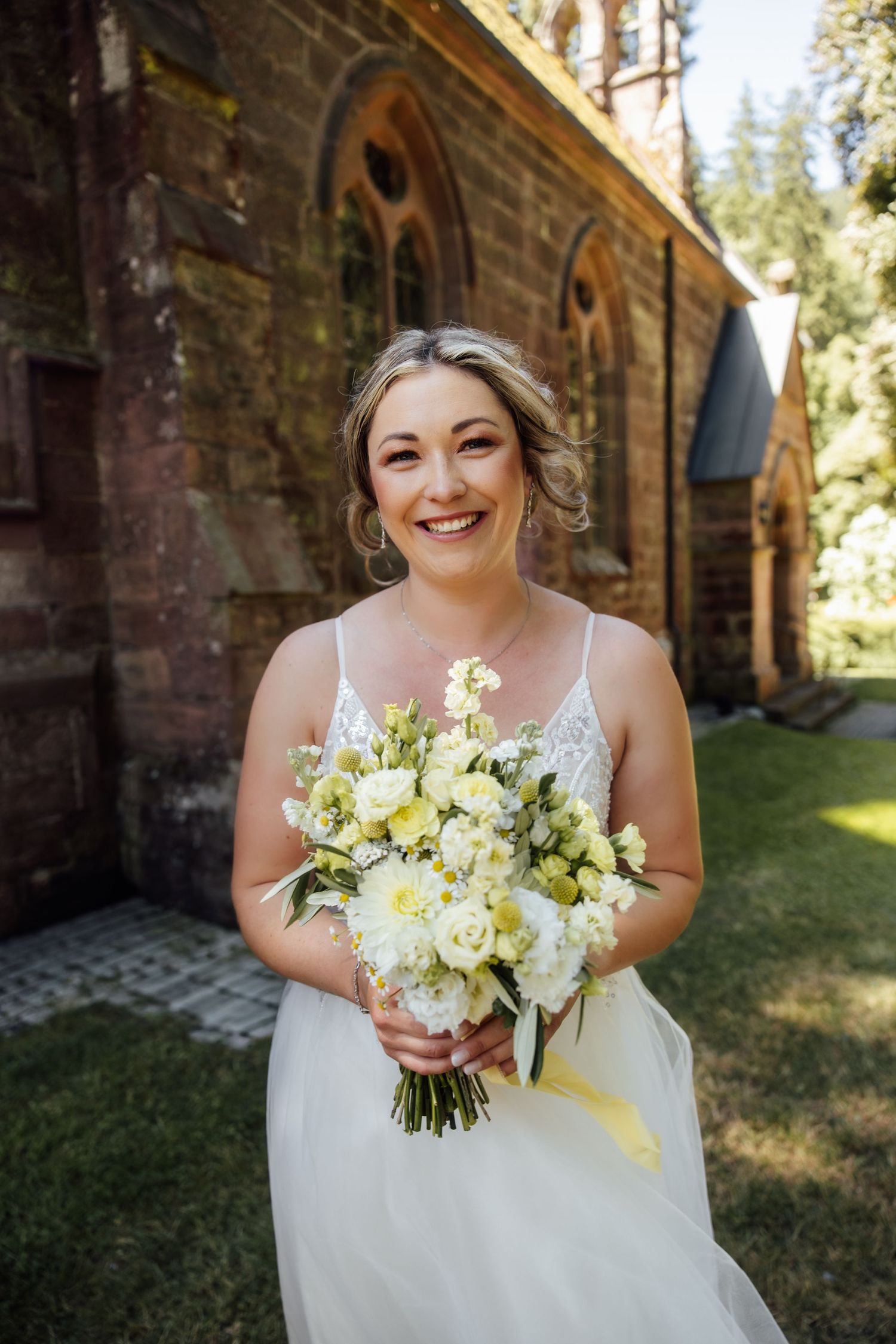 Smiling bride in white gown holds yellow and white bouquet outside a historic brick church on her wedding day.