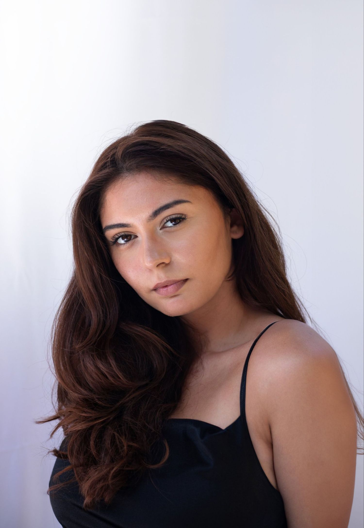 Young woman with long dark hair wearing a black spaghetti strap top posing against a white wall.