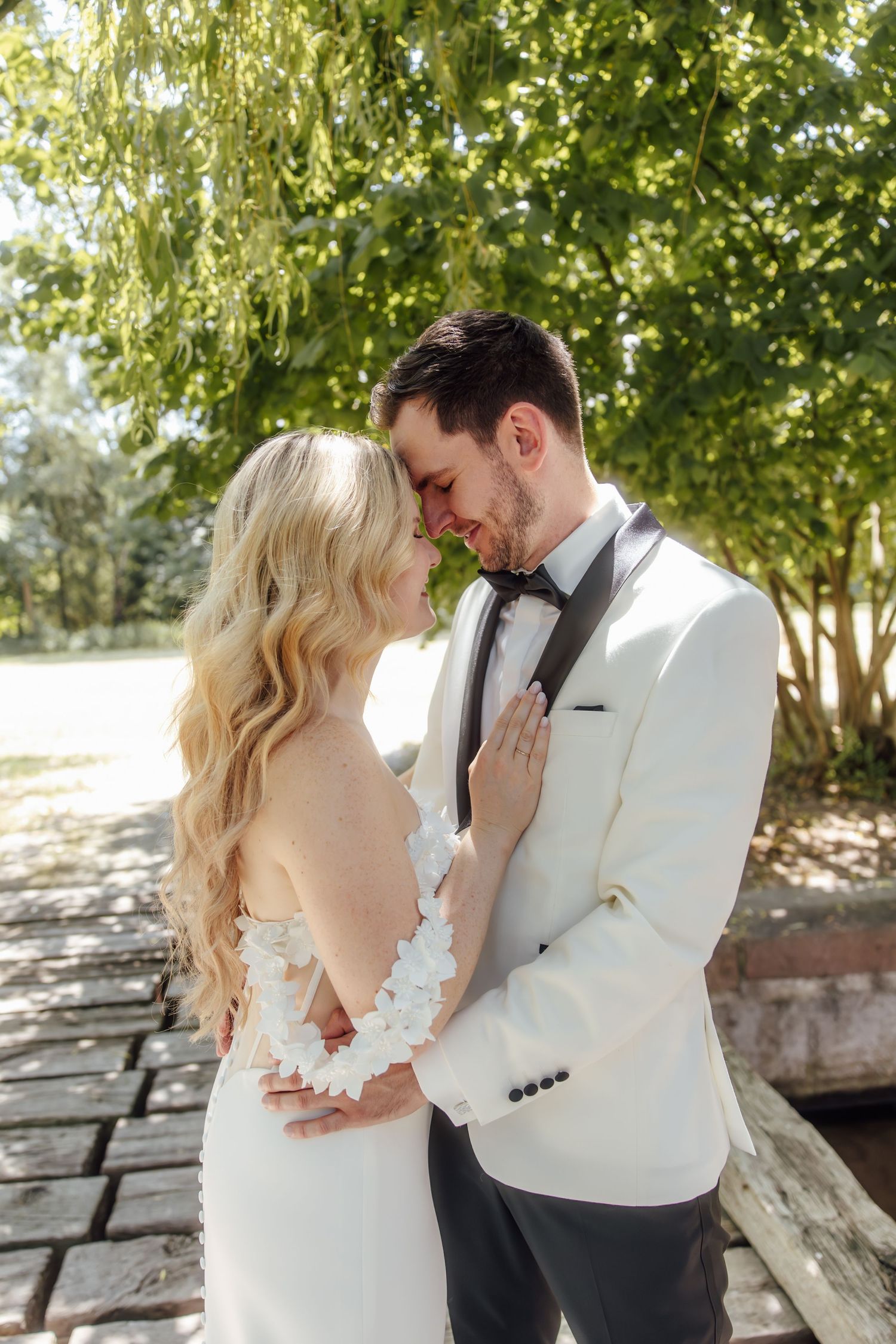 Bride and groom share a romantic moment outdoors, with bride in lace wedding gown and groom in white tuxedo.