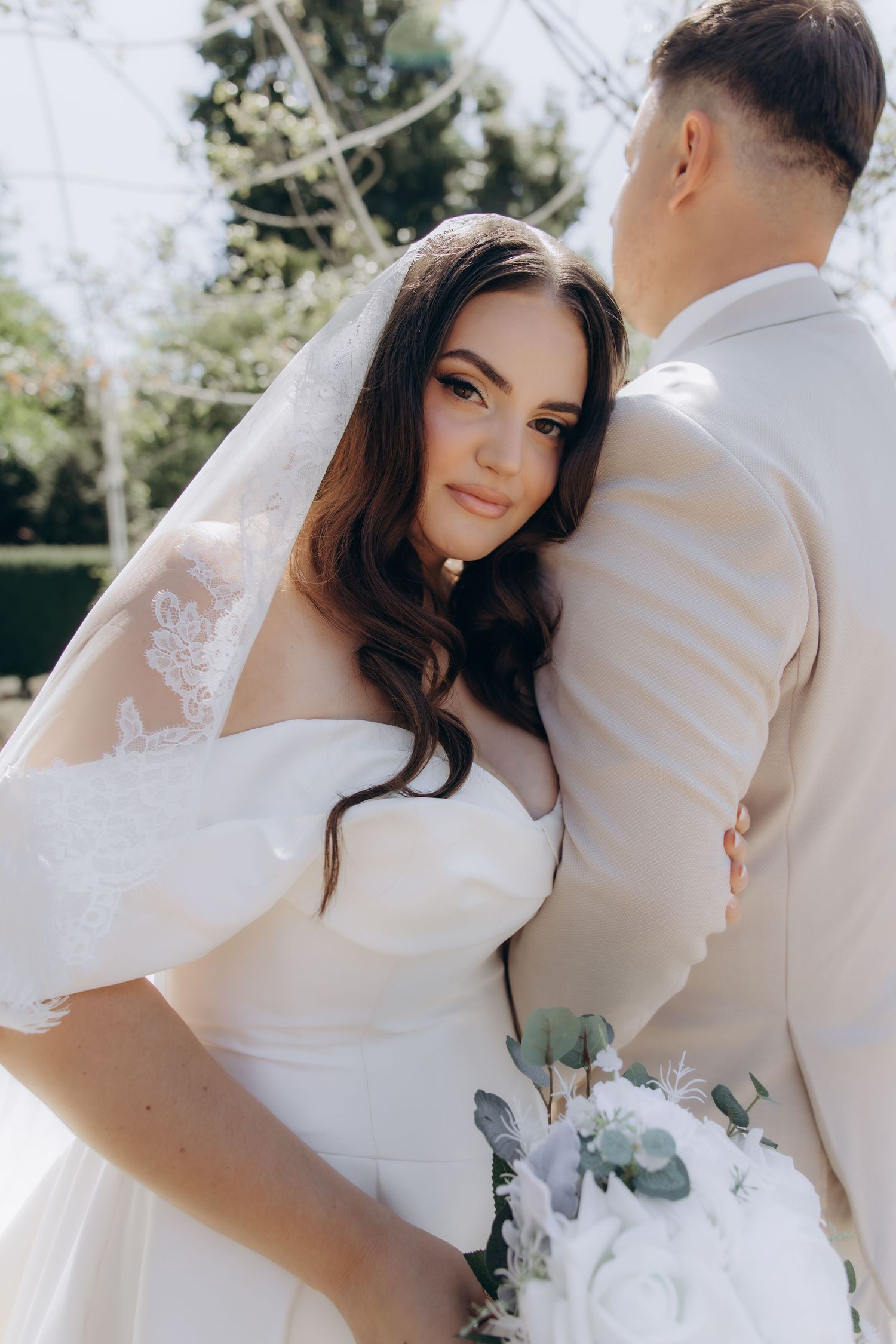 Bride in white gown and veil embraces groom in beige suit outdoors, holding bouquet with eucalyptus.