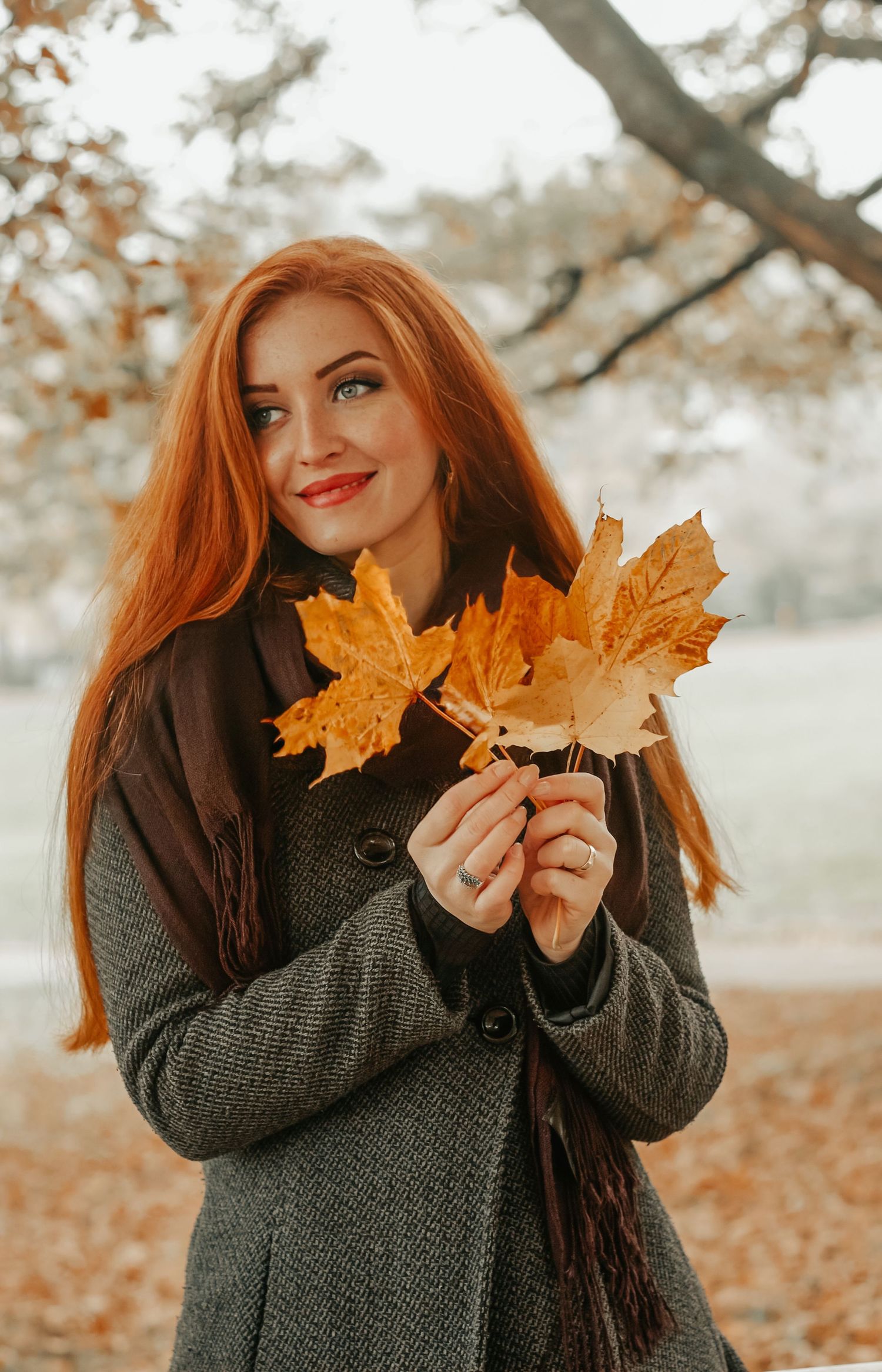 Smiling redhead woman holding autumn maple leaves while standing in a park during fall season.