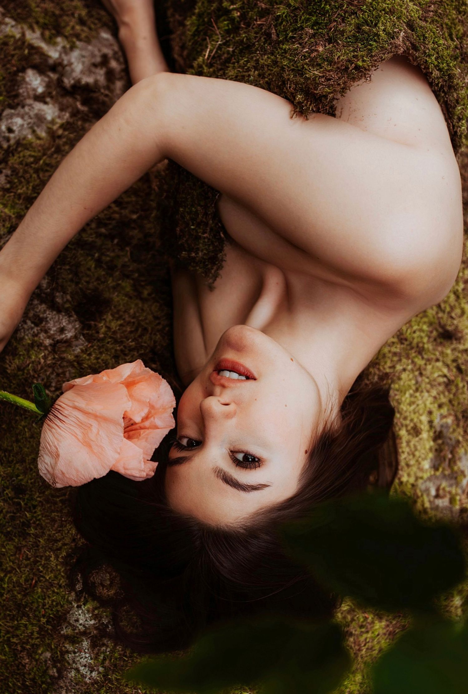 A woman lying on moss holding a pink flower in an artistic nature-inspired fine art portrait.