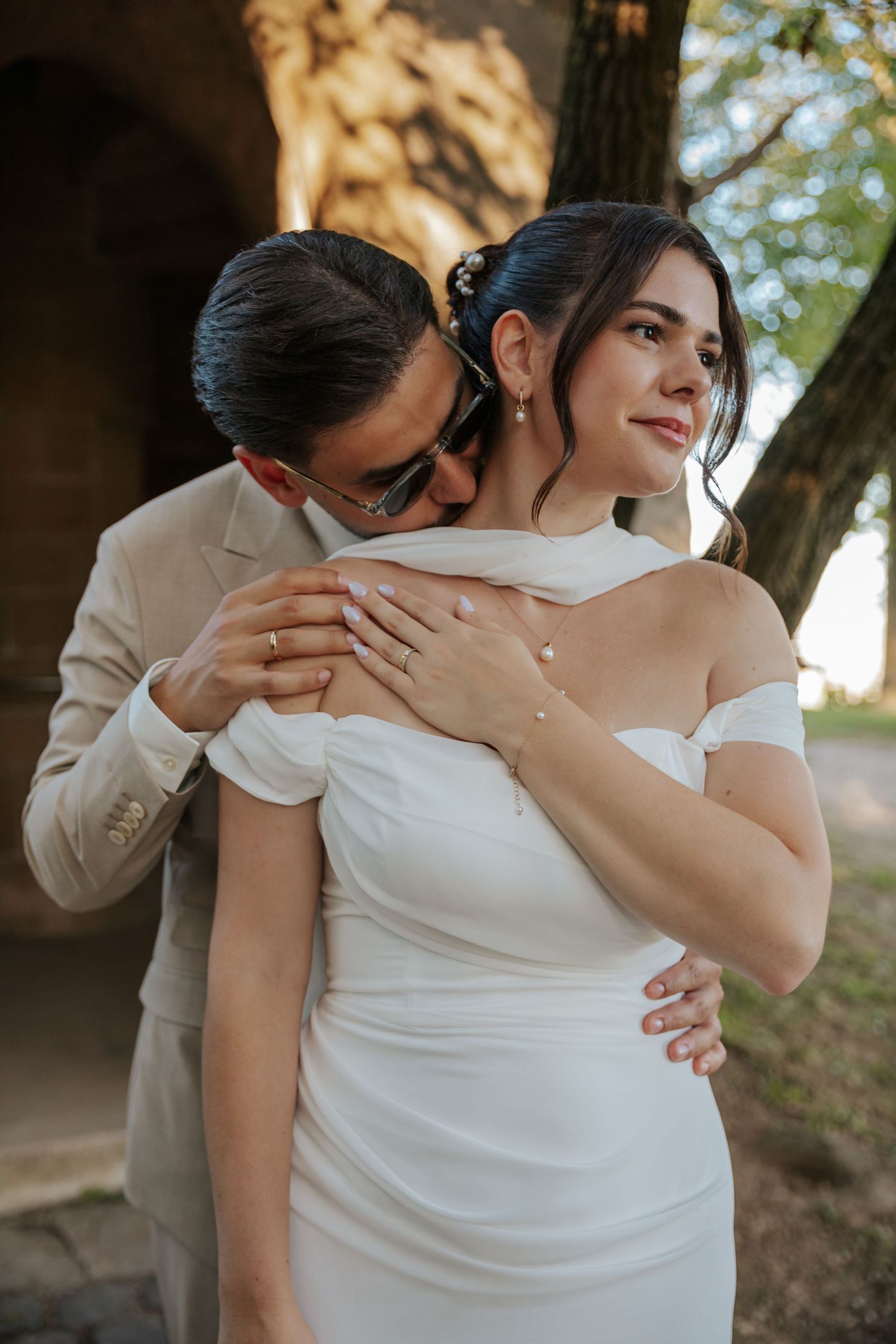 Groom in beige suit embracing bride in off-shoulder white gown from behind near rustic stone arch.
