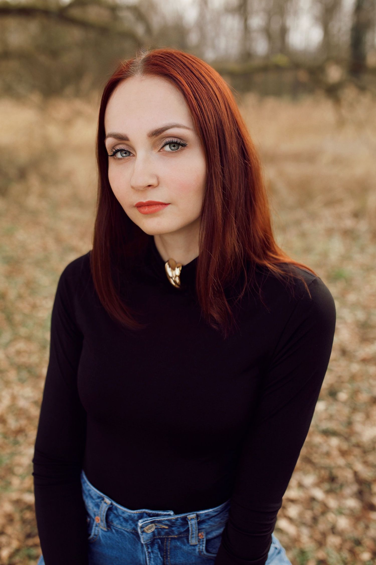 A red-haired woman in a black turtleneck with a gold necklace posing outdoors in autumn.