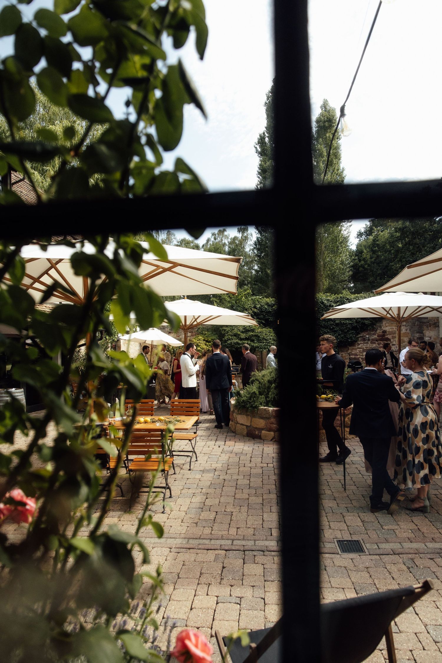 Outdoor garden party with guests mingling under white umbrellas on a brick patio, viewed through a window with lush greenery.