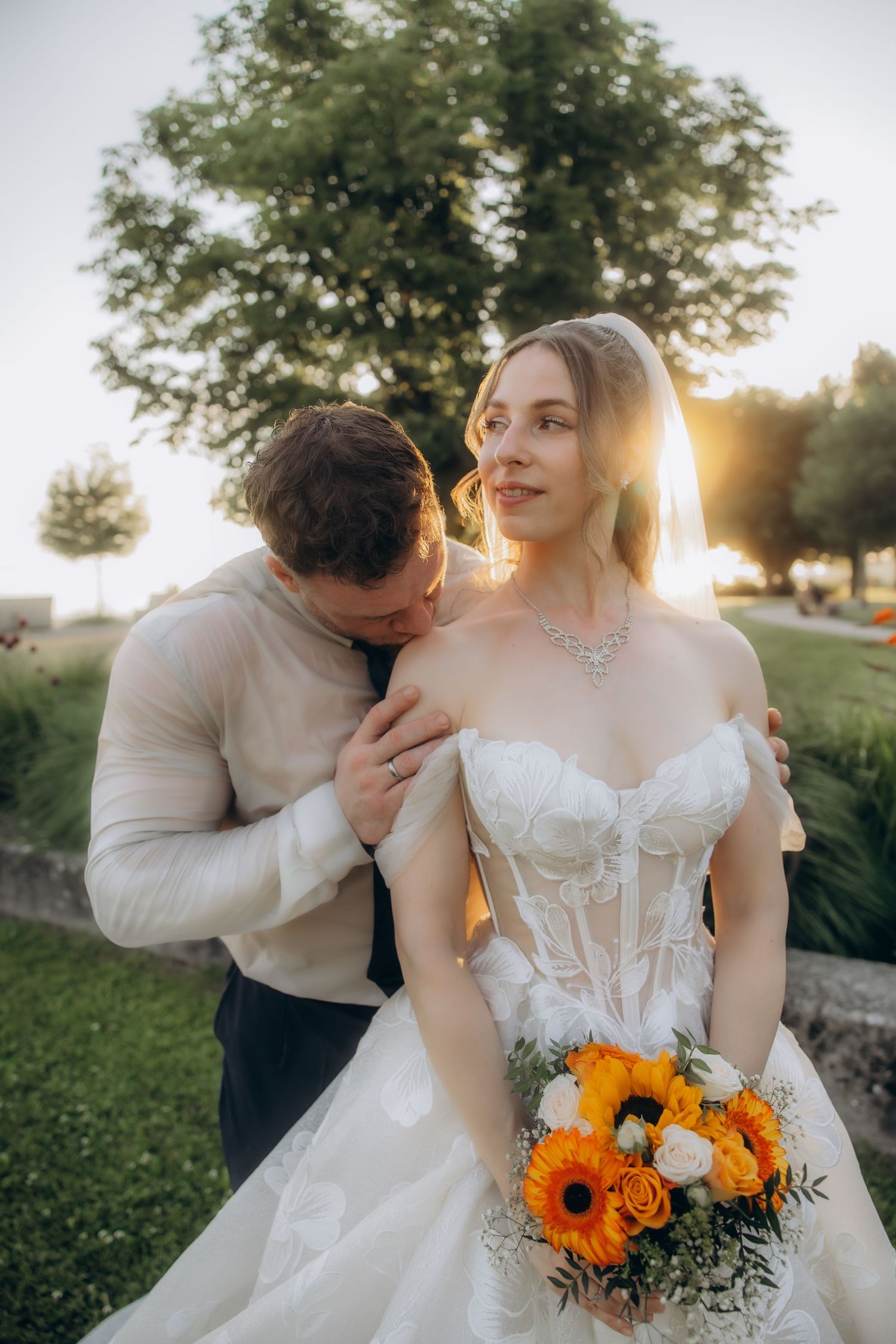 Bride in white off-shoulder gown holds orange bouquet as groom kisses her shoulder at golden hour outdoor wedding.