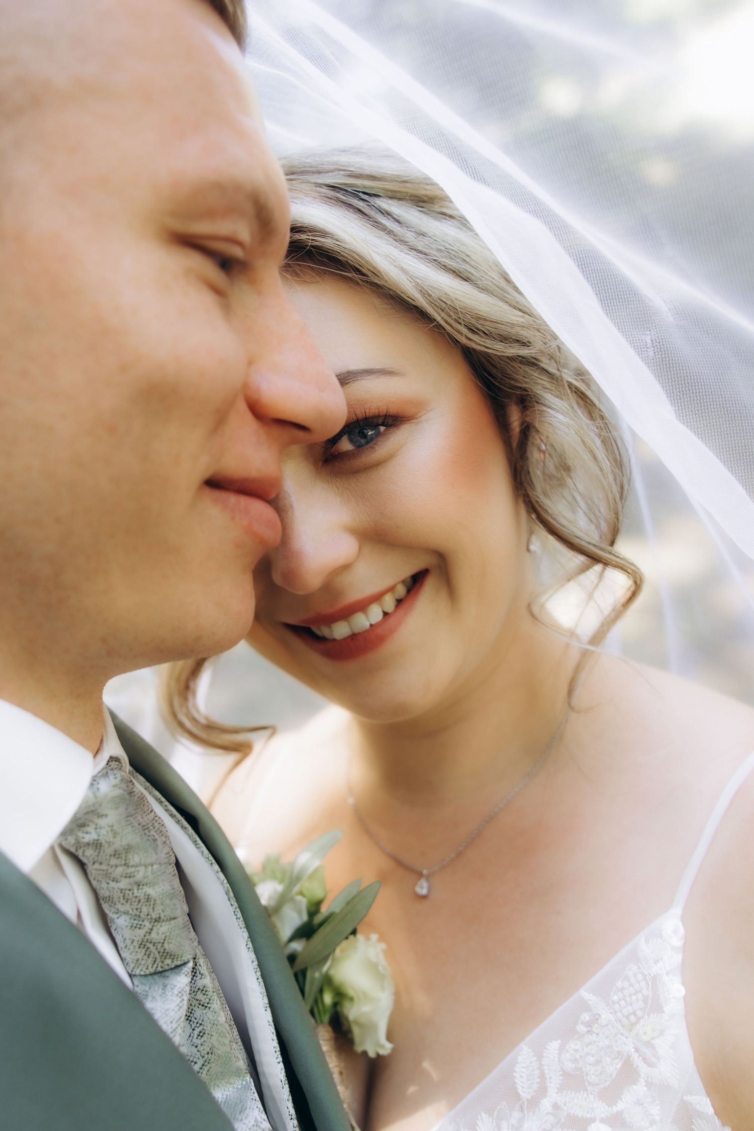 A smiling bride in a lace gown and veil shares a tender moment with her groom in a gray suit with boutonniere.