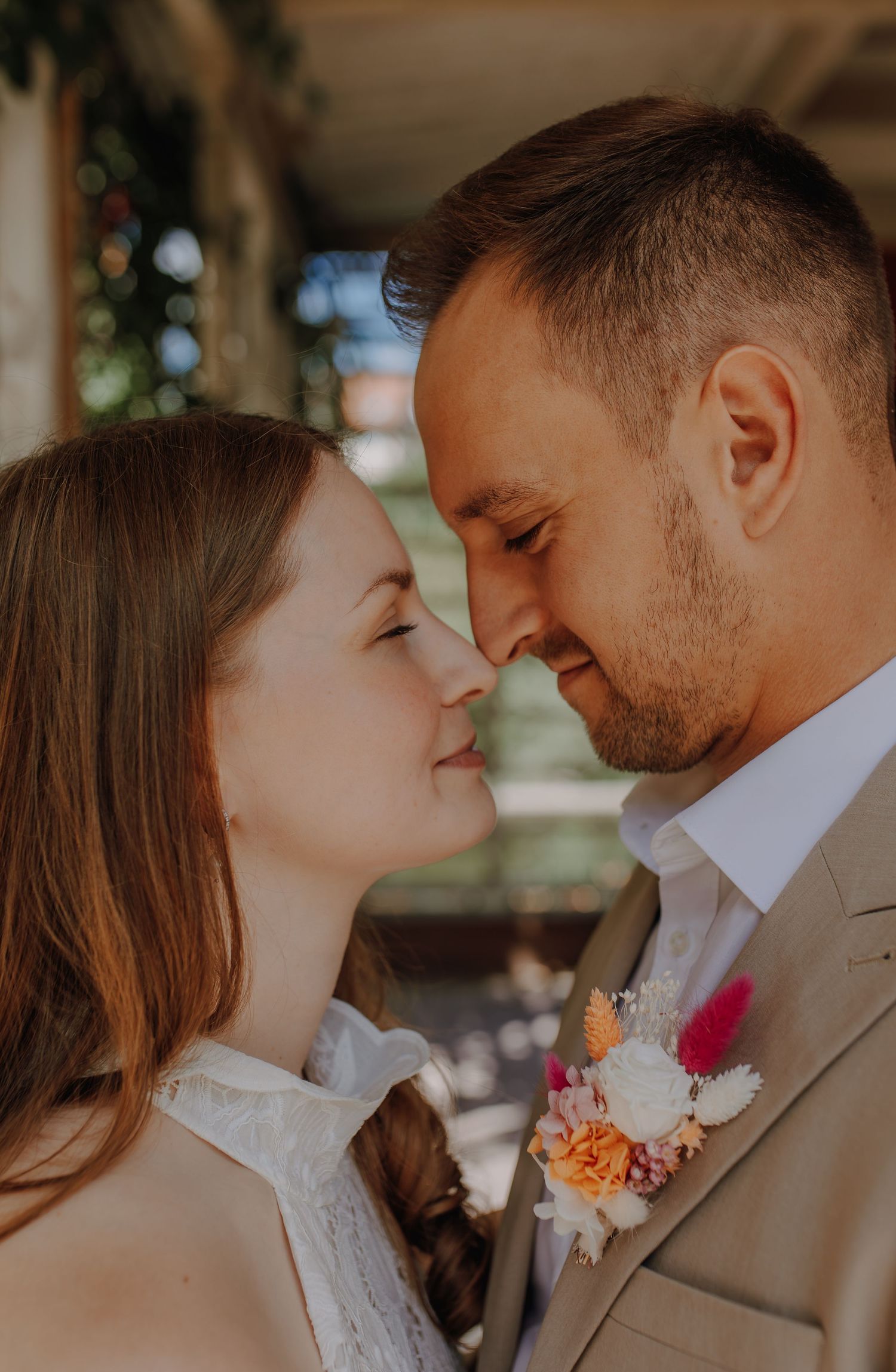 Bride and groom share a tender nose-to-nose moment, groom in beige suit with a colorful floral boutonniere.