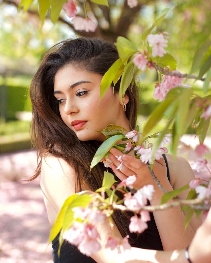 A brunette woman with red lips gazes through pink cherry blossom branches in a sunny garden.