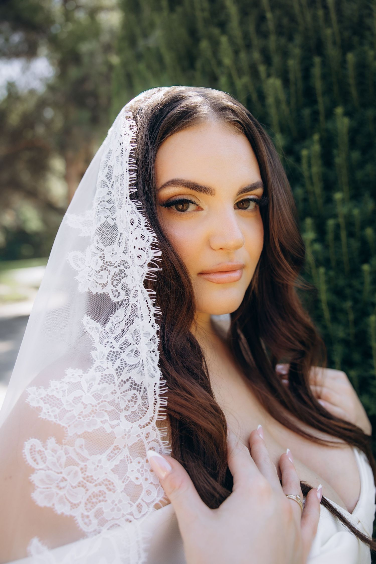 A beautiful bride with dark hair wearing a delicate white lace veil outdoors near green cypress trees.