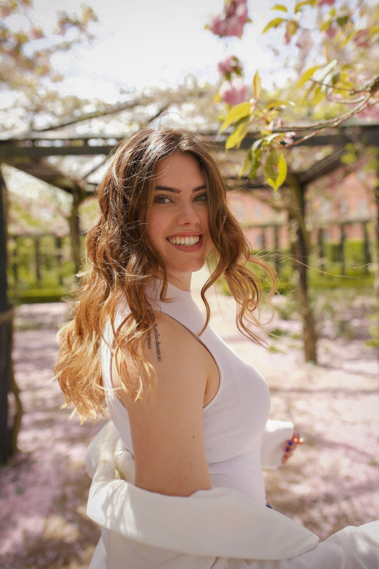 Smiling brunette woman in a white dress spinning outdoors under blooming spring cherry blossom trees.