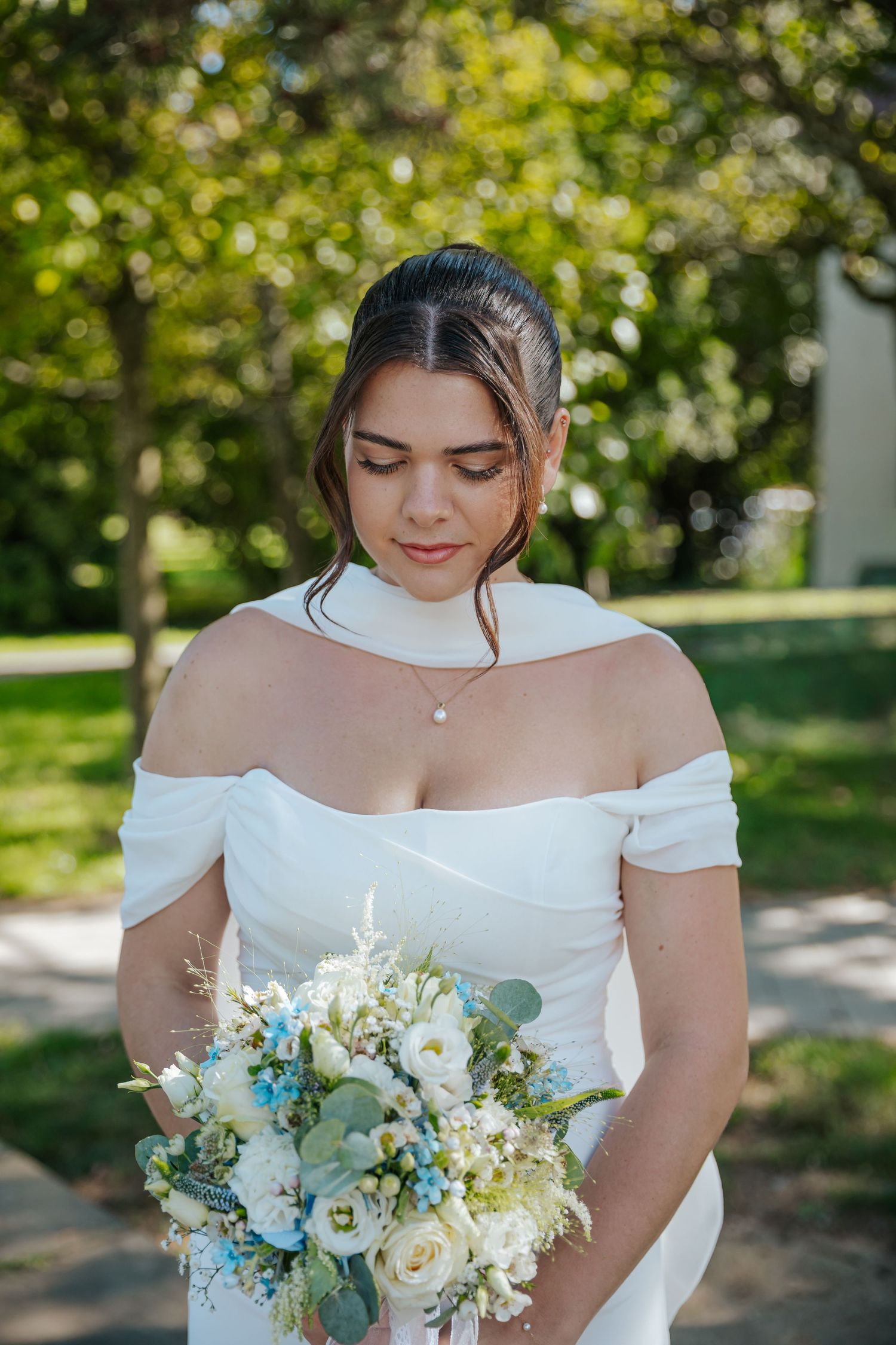 Elegant bride in off-shoulder white gown holding blue and white floral bouquet in sunny park setting.