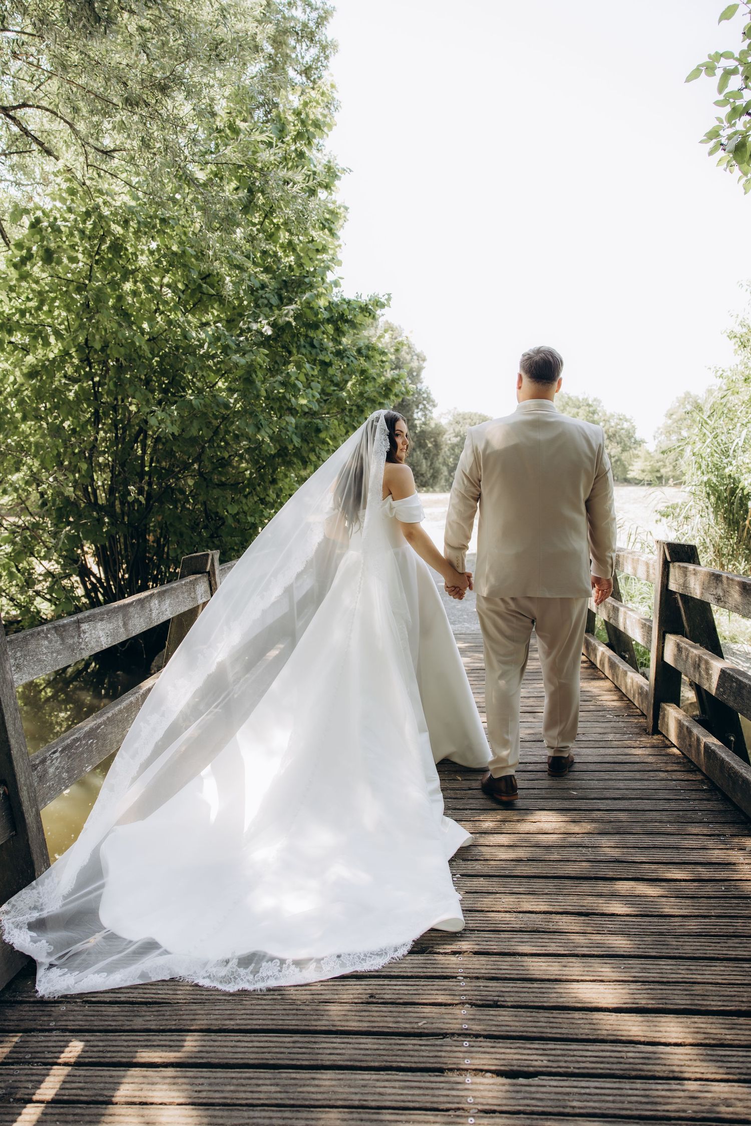 Bride and groom walking hand in hand on a wooden bridge surrounded by lush green trees on their wedding day.