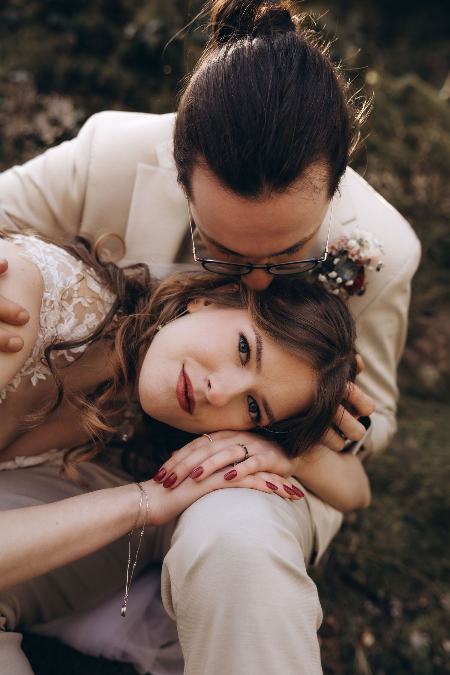 Groom in white suit kissing bride on head as she rests against him outdoors.