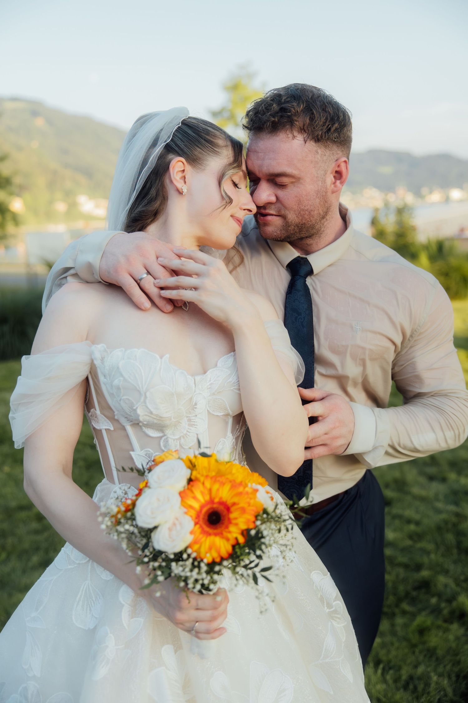 Bride and groom share a romantic moment outdoors, bride holding orange and white bouquet, mountains in background.