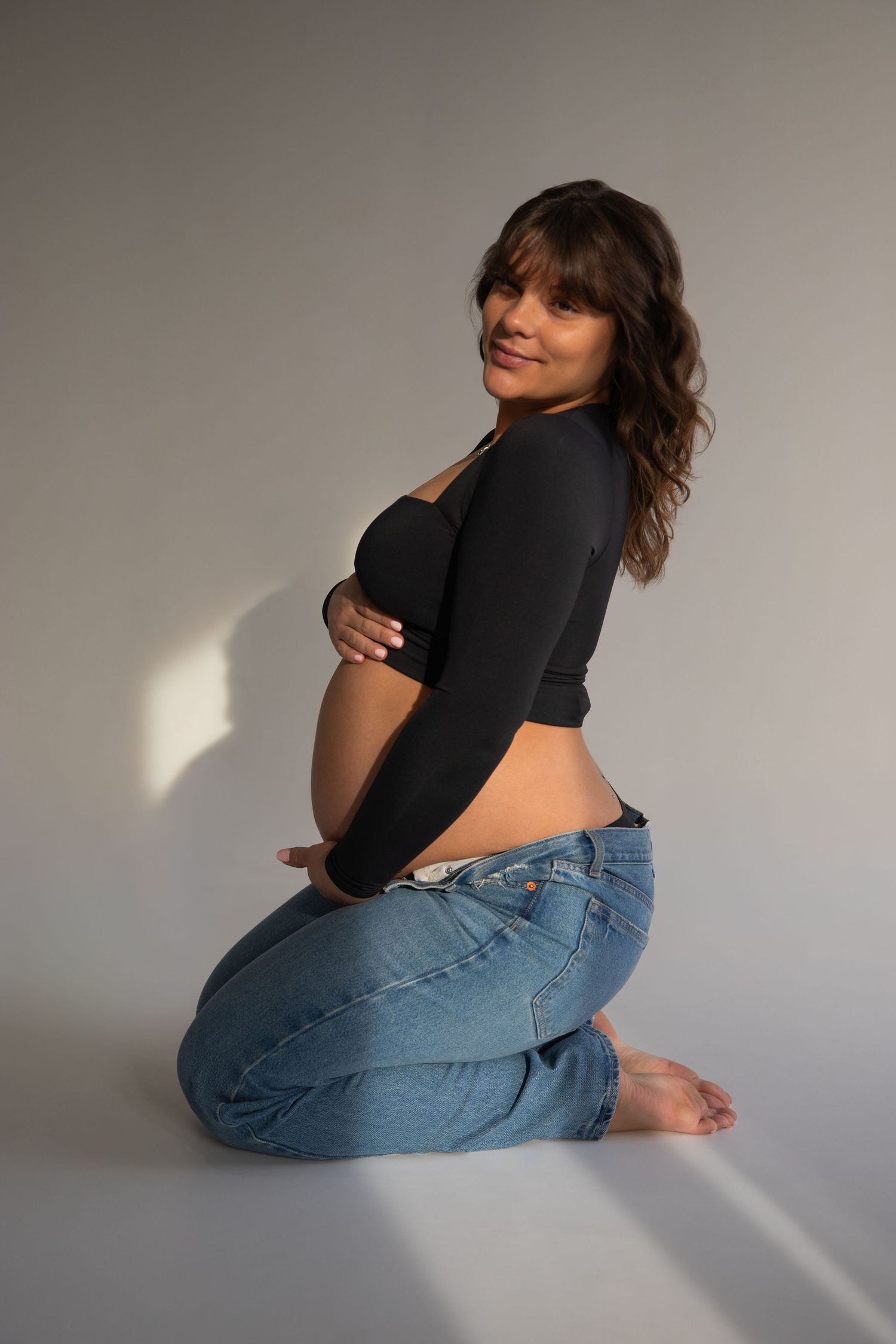 A smiling pregnant woman kneeling barefoot on a studio floor wearing a black crop top and blue jeans.