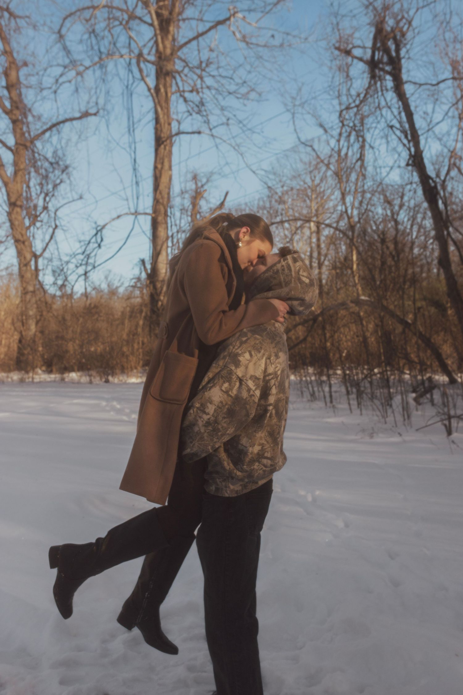 Couple embracing and kissing in a snowy winter park, wearing warm coats among bare trees.