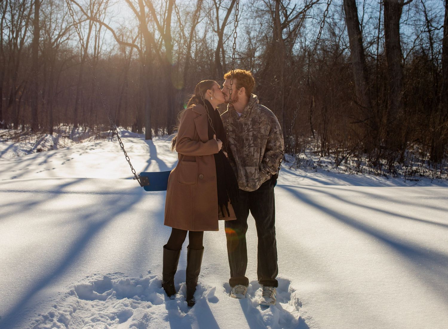 Couple sharing a romantic kiss in a snowy winter forest with sunlight streaming through bare trees.