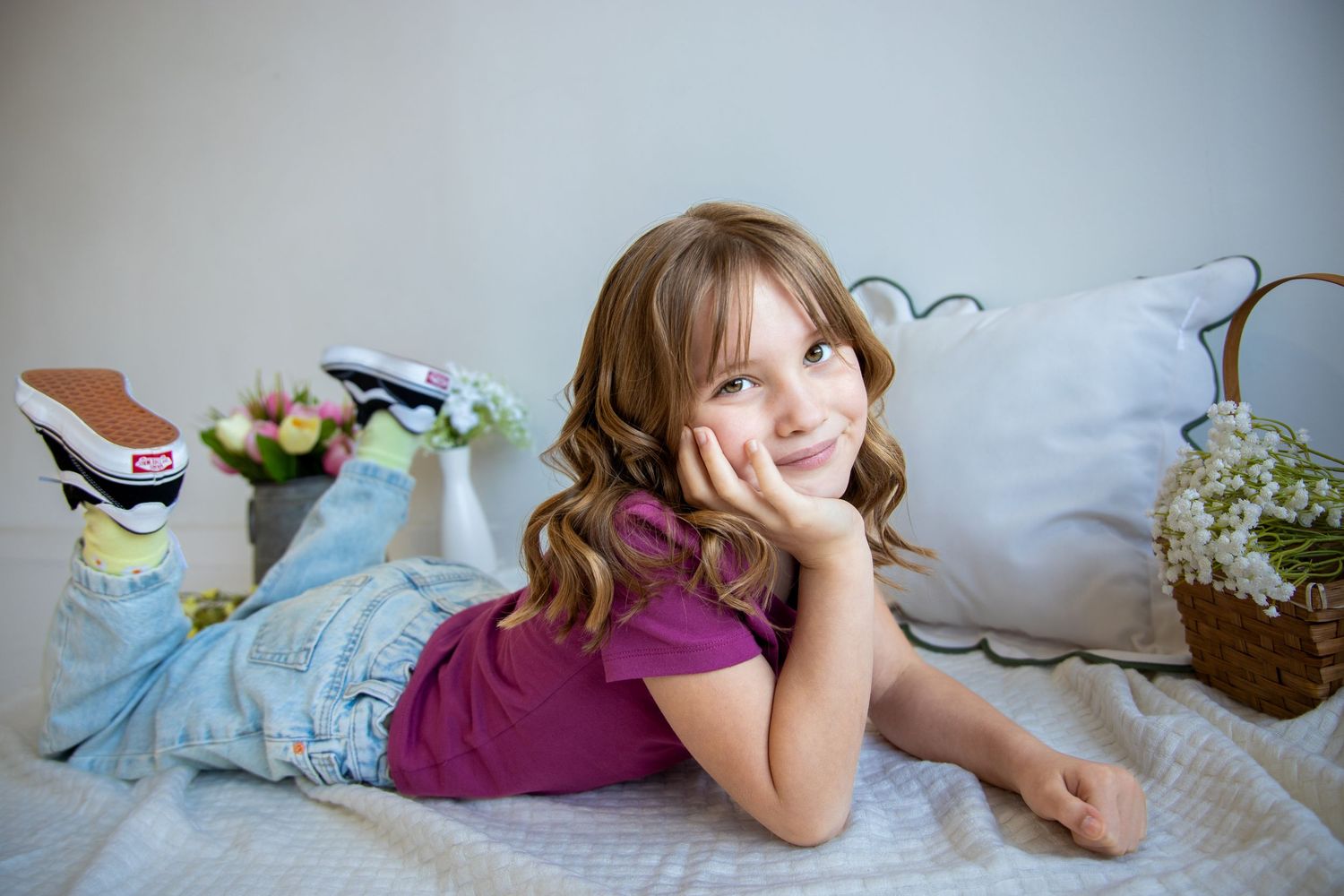 A happy young girl with curly hair posing on a white bed wearing a purple top and blue jeans.