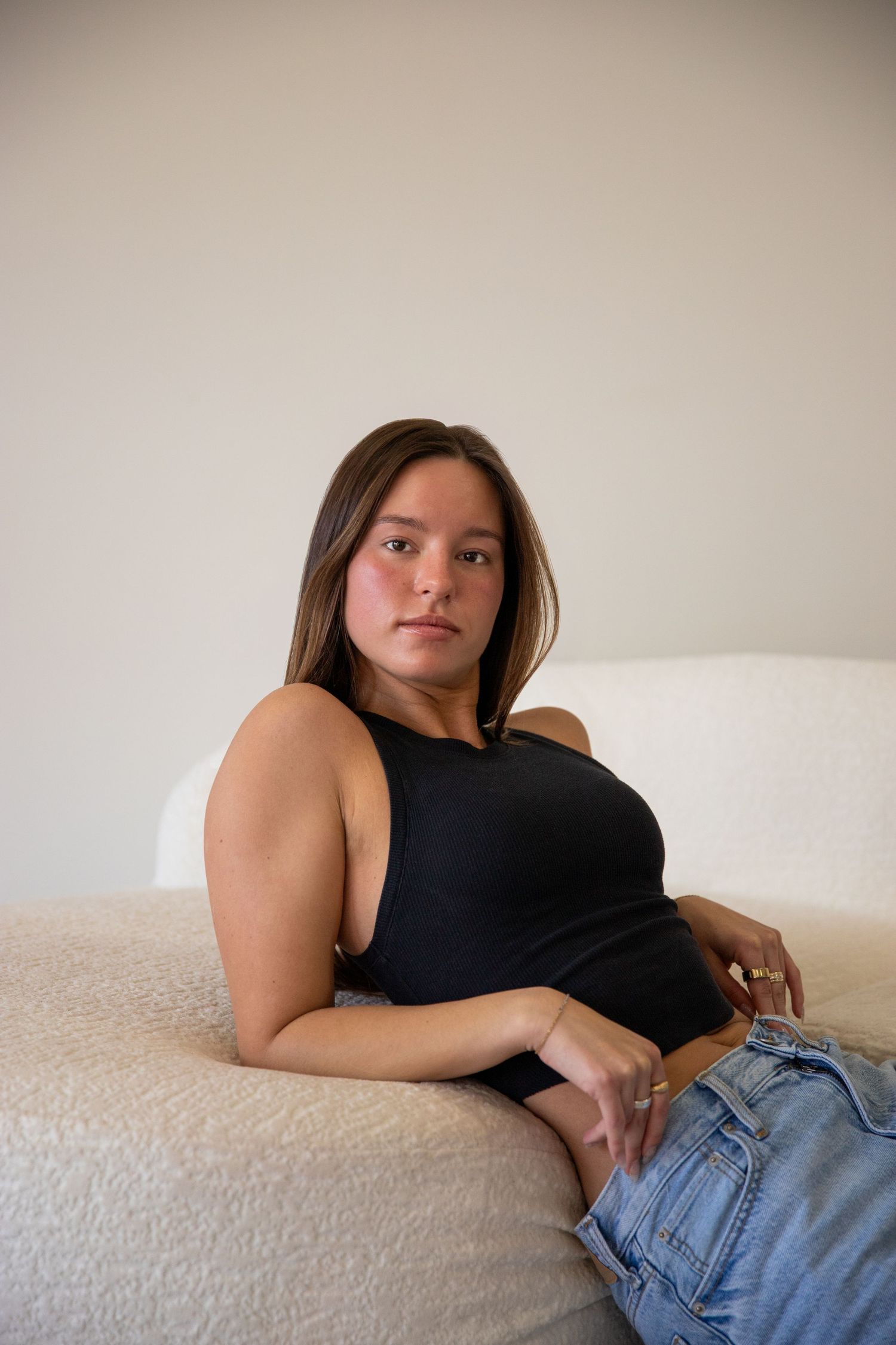 Young woman in black tank top and jeans sitting relaxed on a white couch looking at camera.