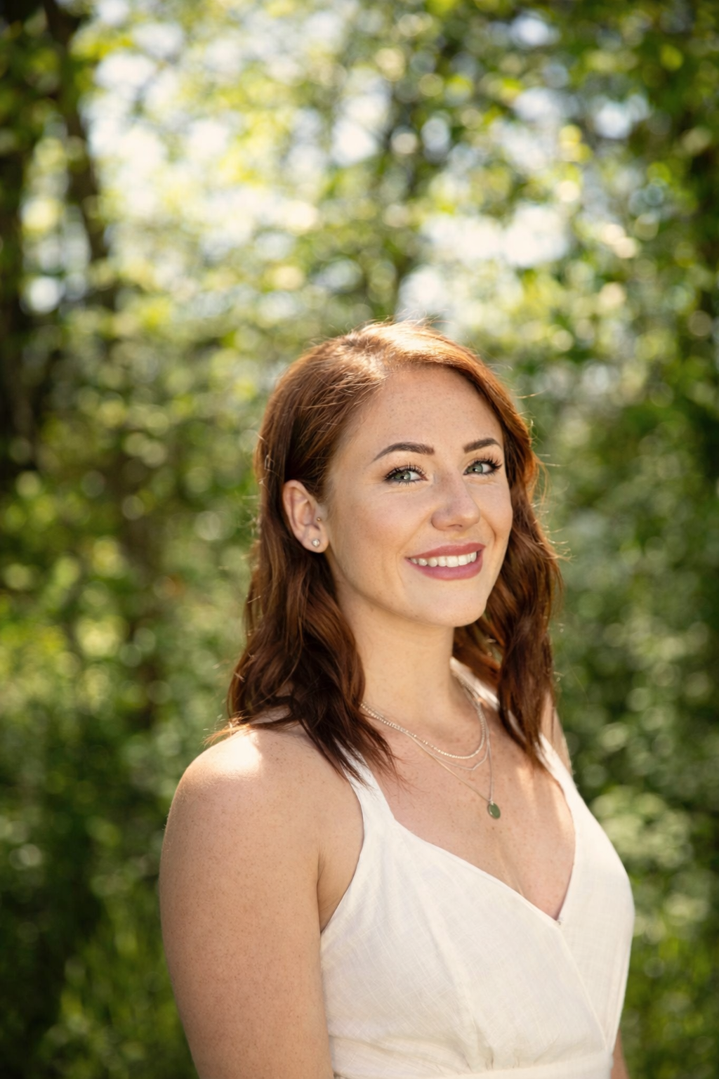 Smiling young woman with red hair in a white dress posing outdoors among lush green trees.