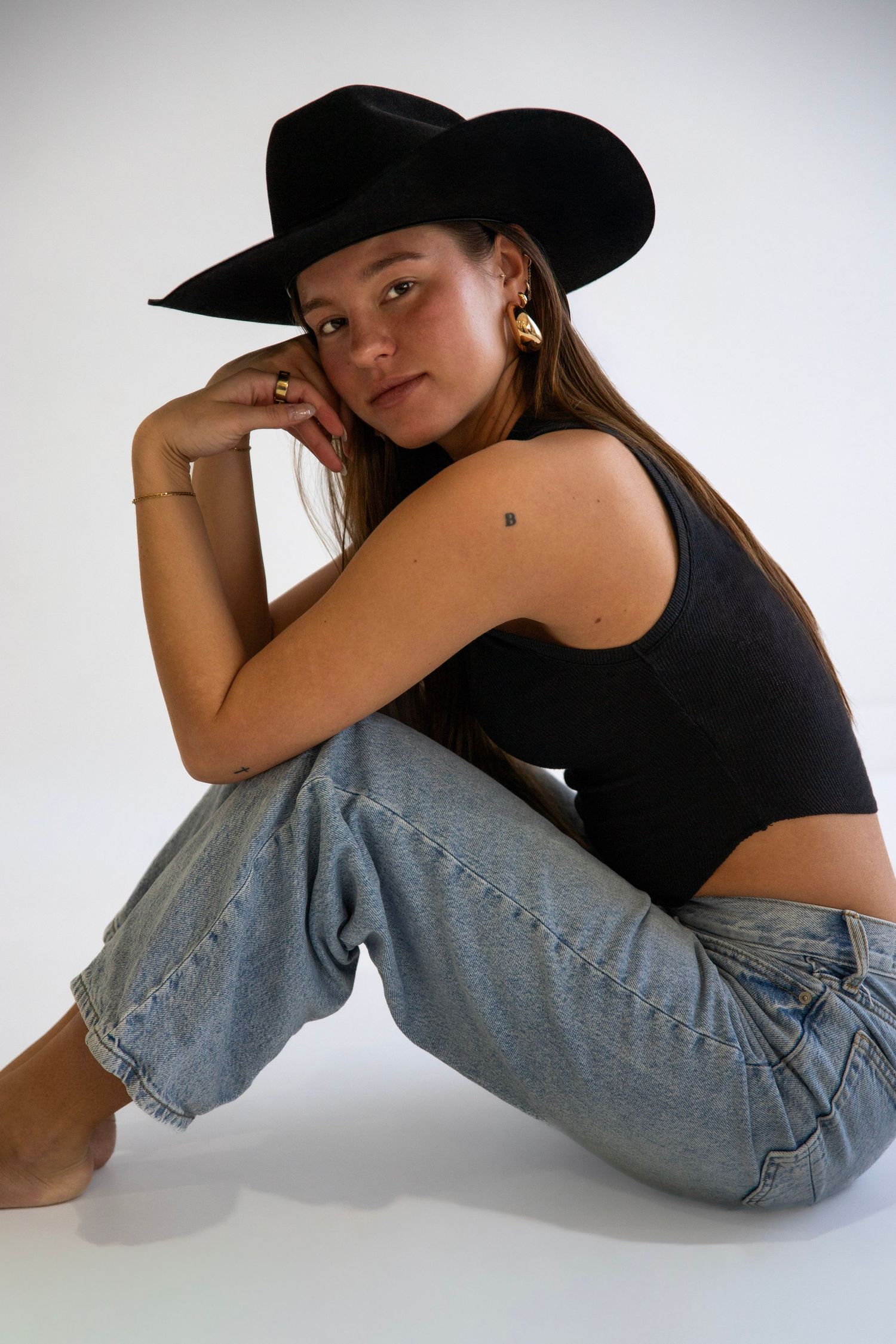 Young woman in black crop top, jeans, and black cowboy hat sitting on floor in studio.