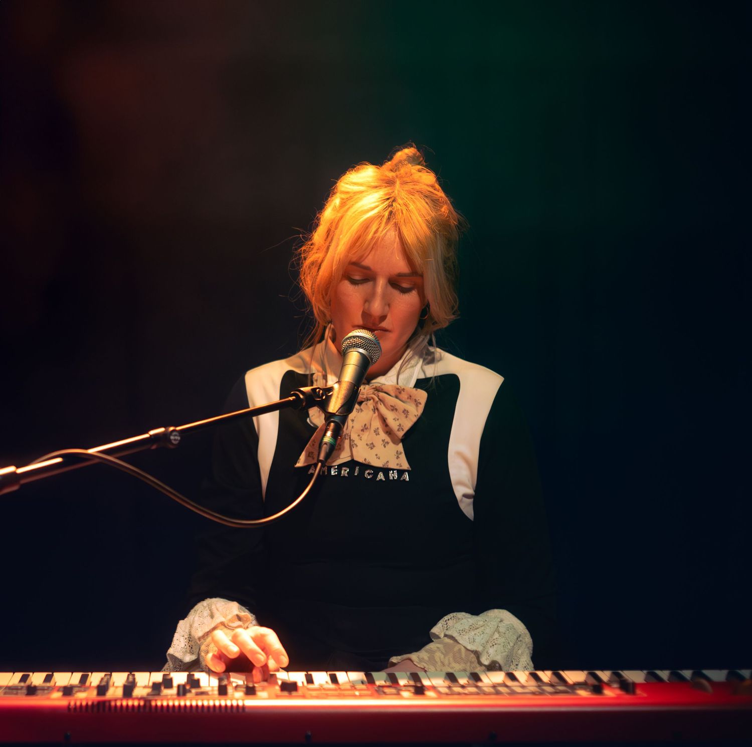 Blonde woman playing keyboard synthesizer and singing into microphone on stage under dramatic lighting.