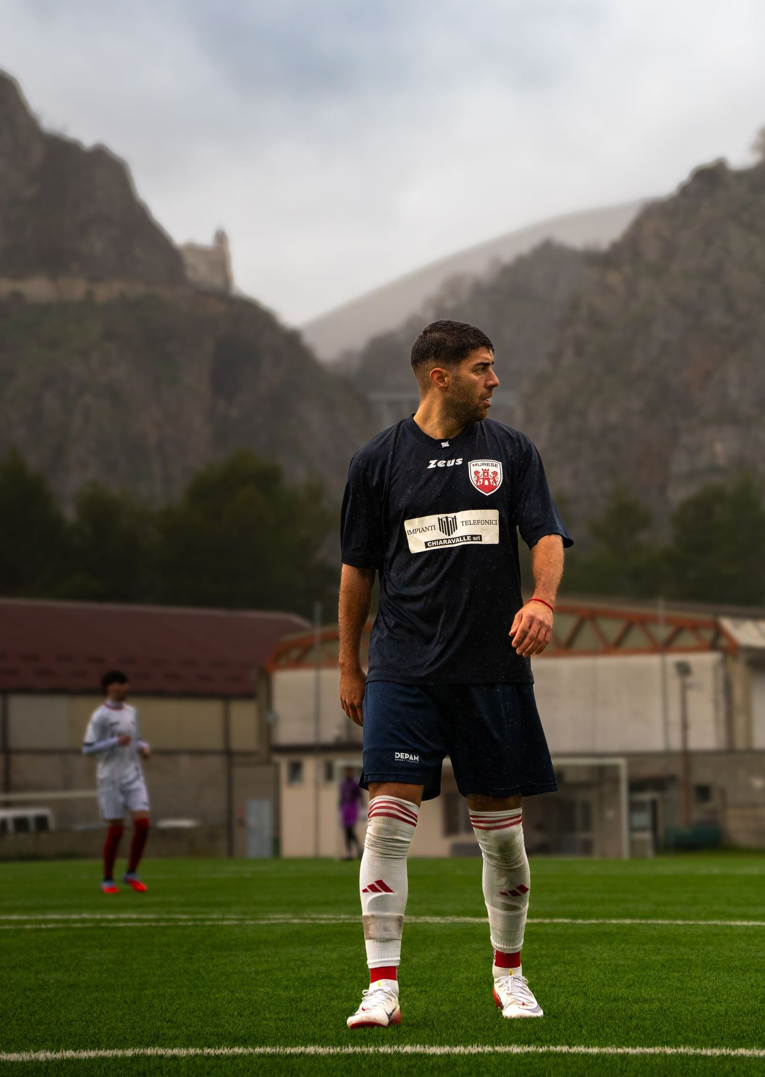 A young soccer player in a navy blue uniform stands on a green field with dramatic rocky mountains in the background.