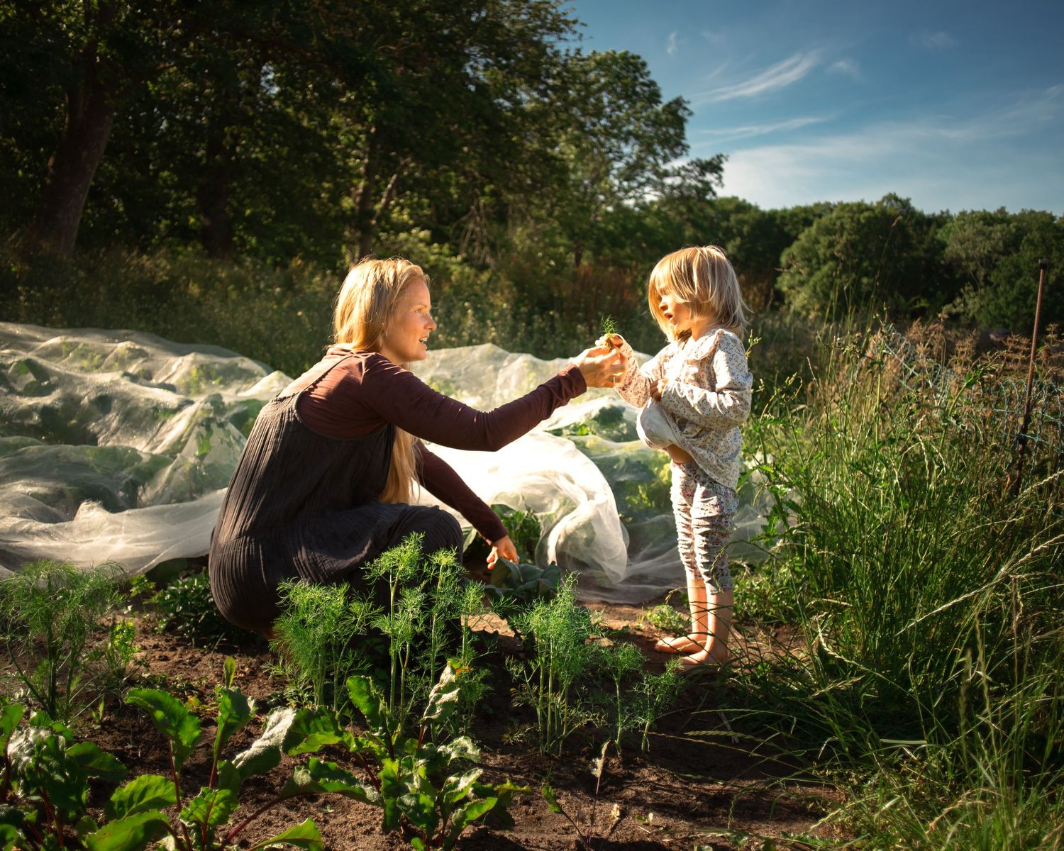 Woman kneeling in garden sharing vegetables with young child on sunny day surrounded by lush greenery.