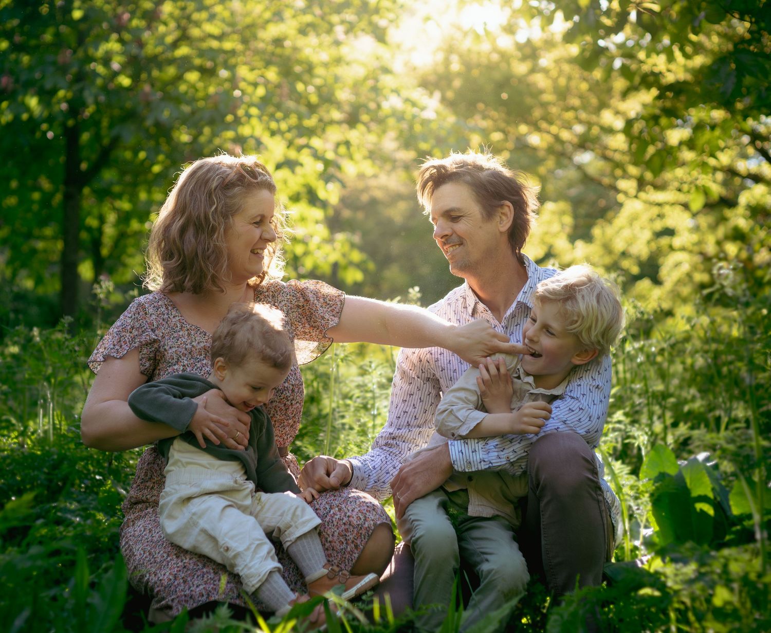 Happy family with two young children sitting together in a sunlit green forest, sharing a joyful moment outdoors.
